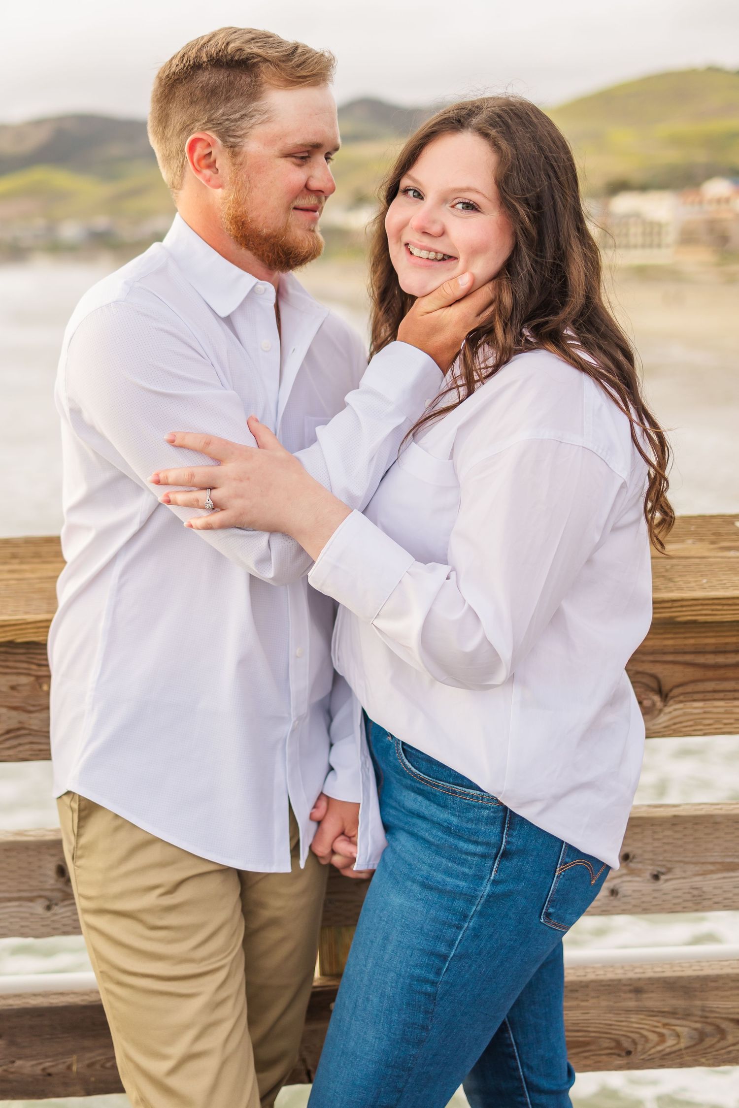 A happy couple embracing and smiling on a wooden pier with mountains in the background.