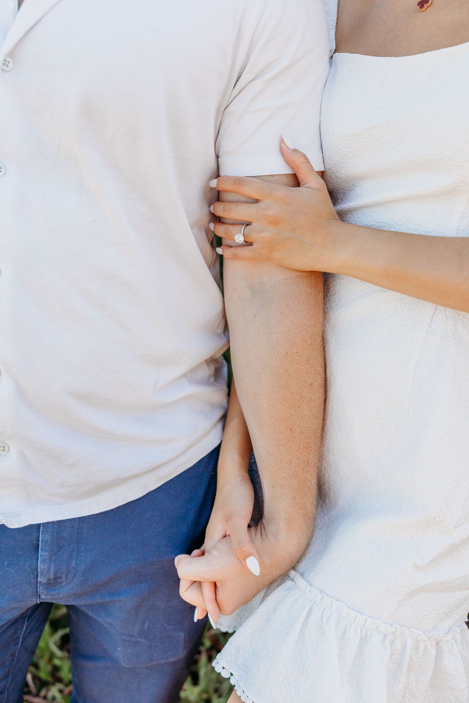 Close-up of intertwined hands showing off an engagement ring against white clothing.