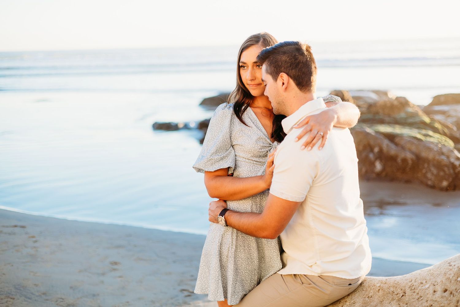 A loving couple holds each other close on a beach with rocky coastline.