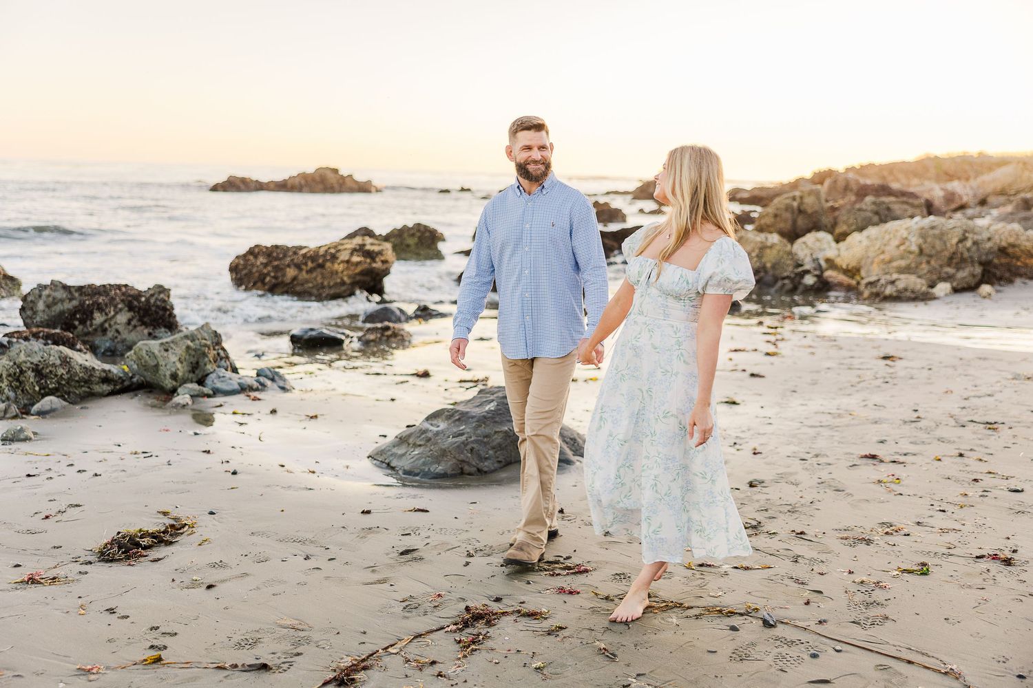 A couple strolls hand in hand along a rocky beach shoreline at sunset.