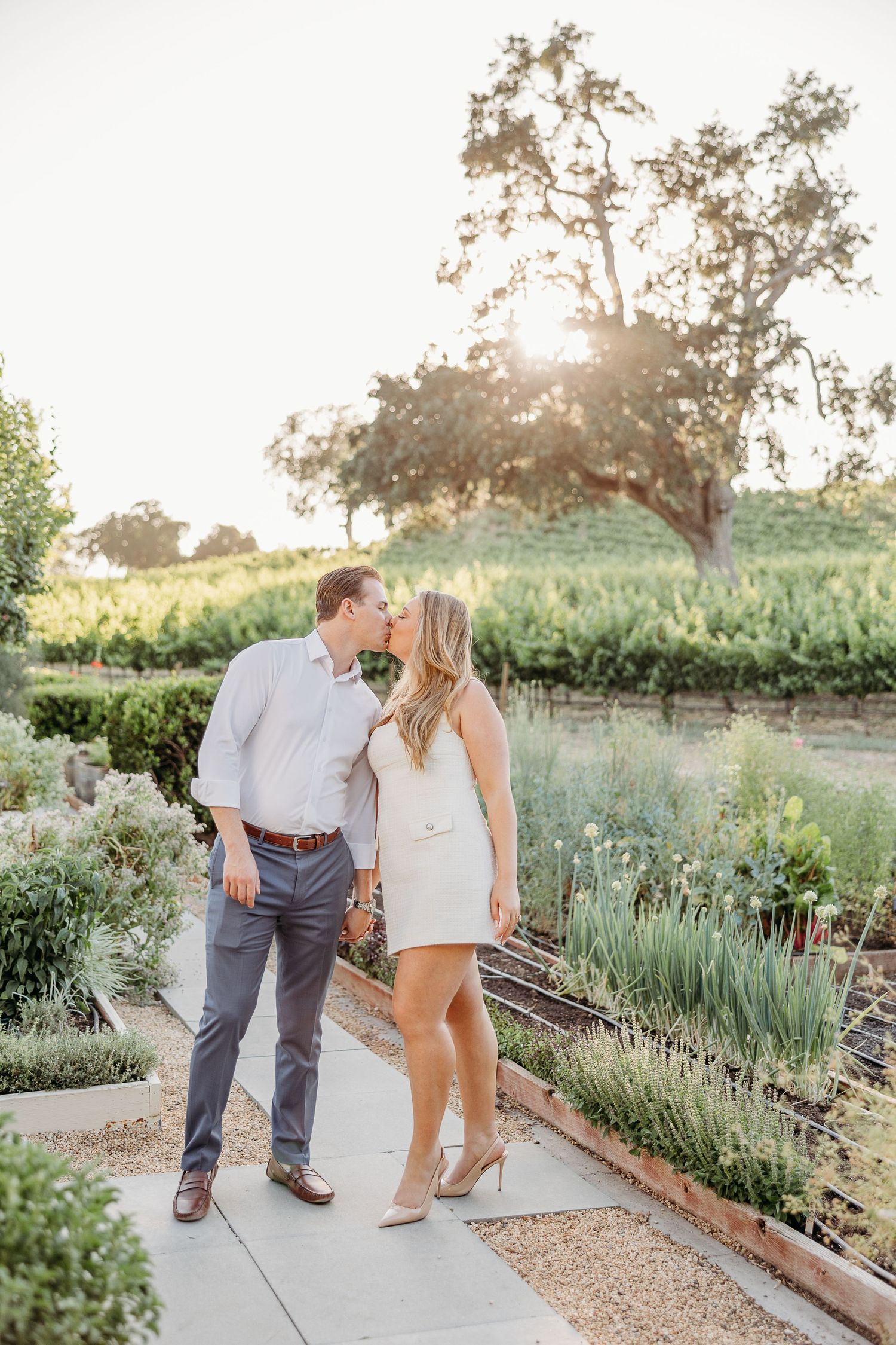 A romantic couple shares an intimate moment while walking along a garden path at sunset.