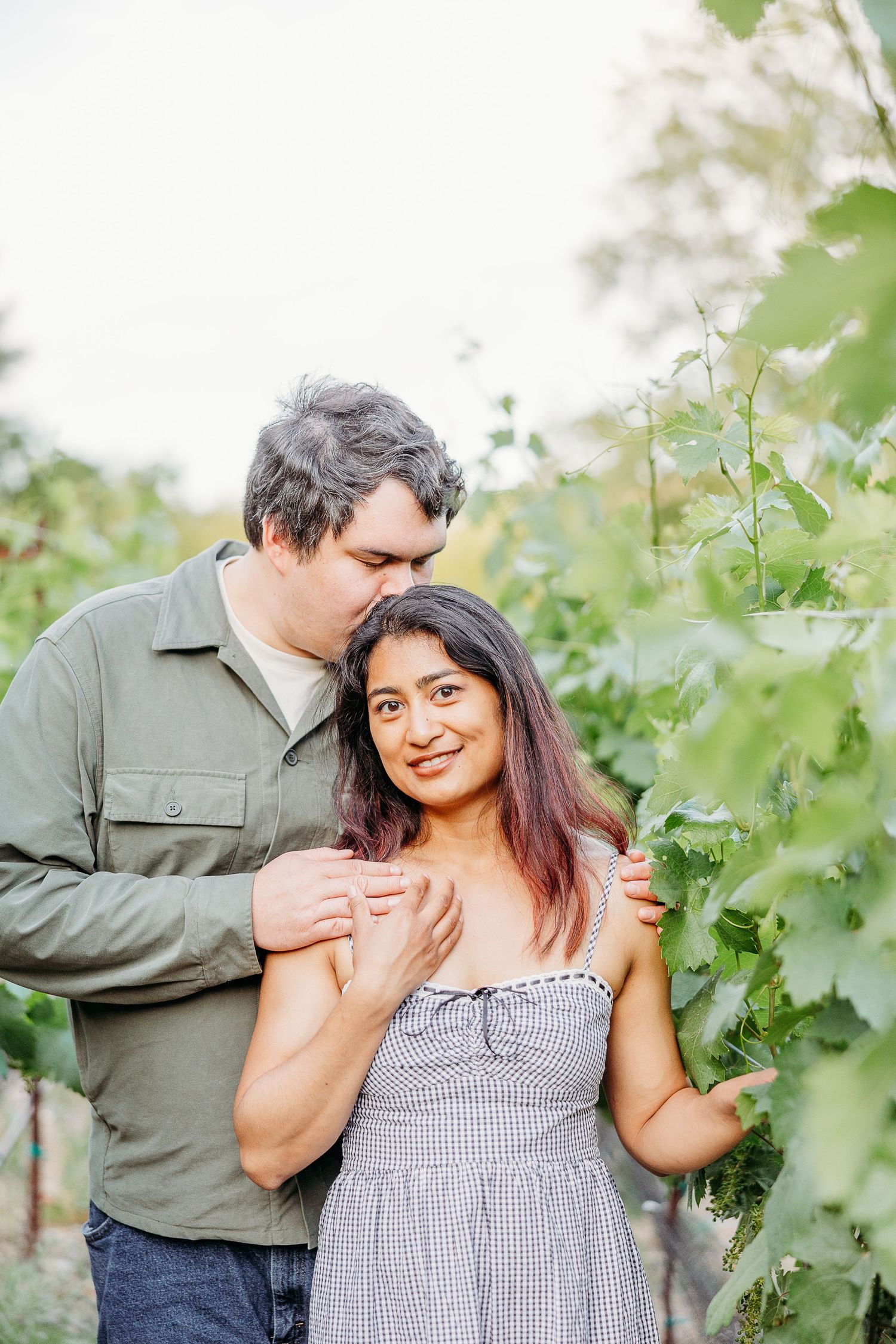 A couple stands together in a vineyard with grapevines on a sunny afternoon wearing casual attire.