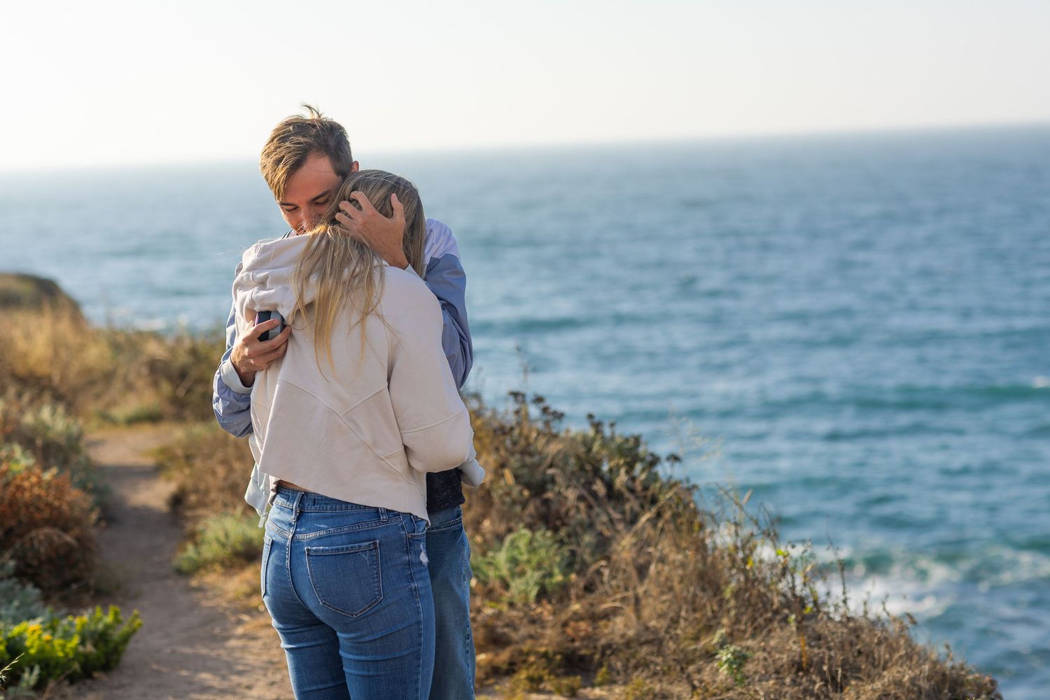 A couple embraces on a coastal path with the blue ocean in the background.