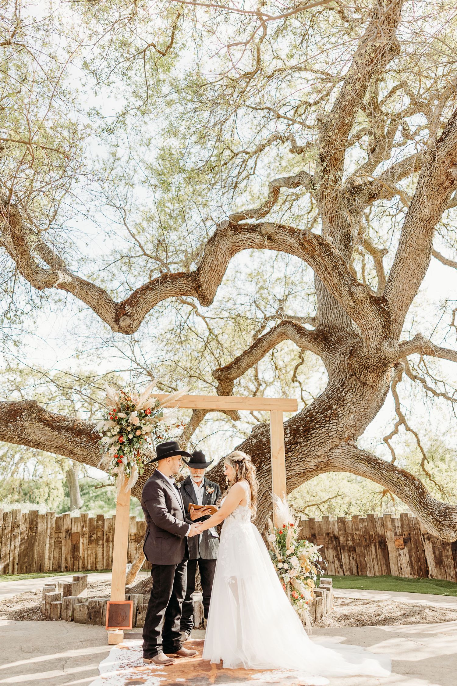 A wedding ceremony under a majestic oak tree with a wooden arch decorated with floral arrangements.