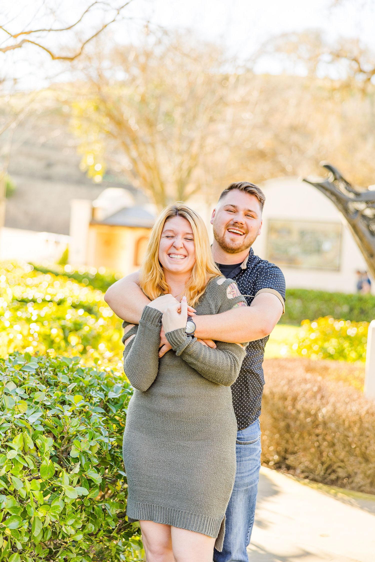 Couple shares playful moment while embracing in front of manicured garden landscape.