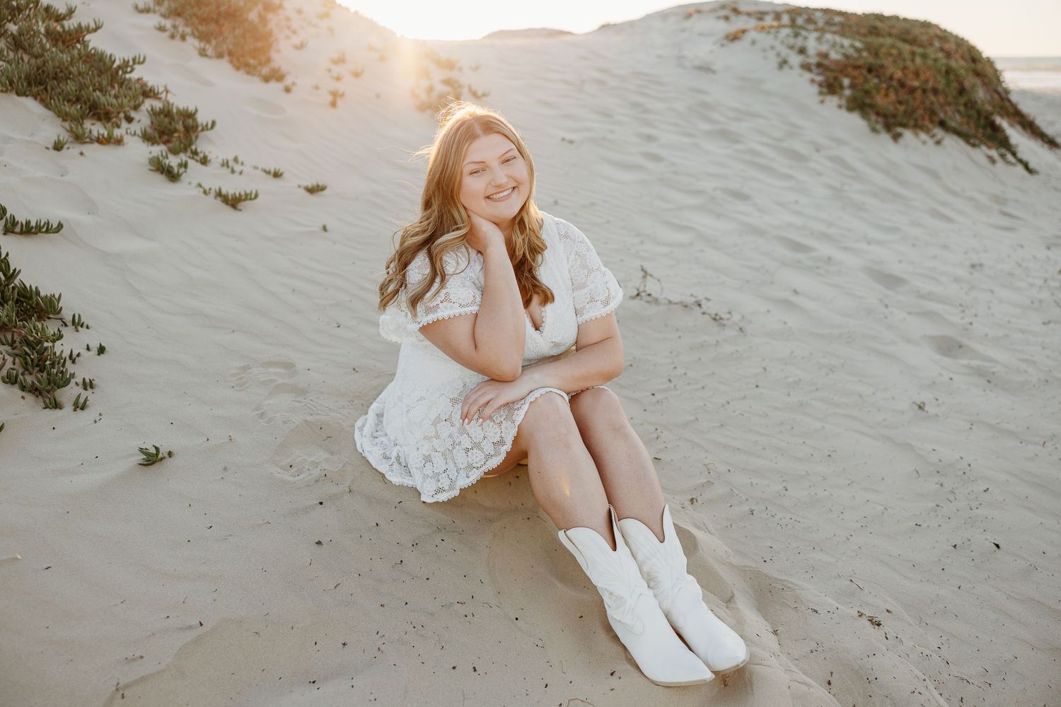 Graduate in white dress and cowboy boots sits on sandy dunes during sunset photo session.