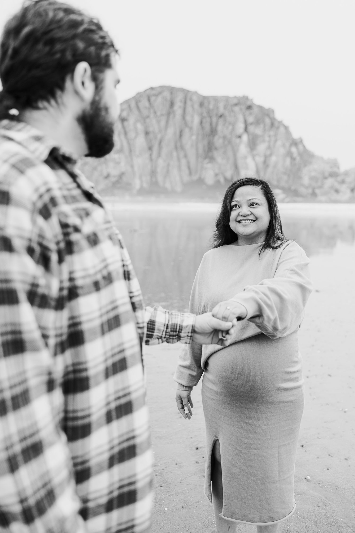 A couple stands on a beach with Morro Rock in the background during a black and white photoshoot.