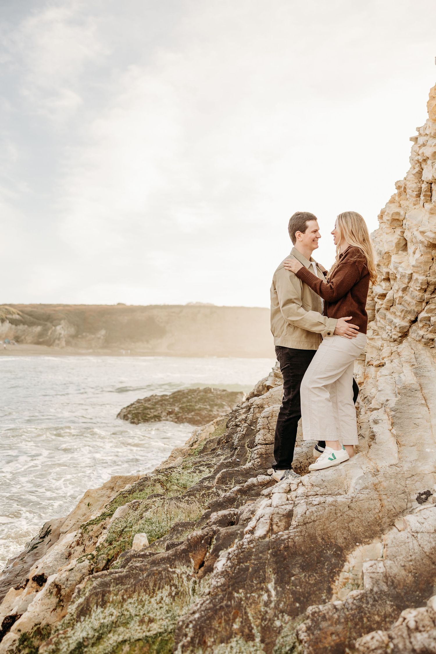 A romantic couple embraces on rocky coastal cliffs overlooking a dramatic ocean landscape at sunset.