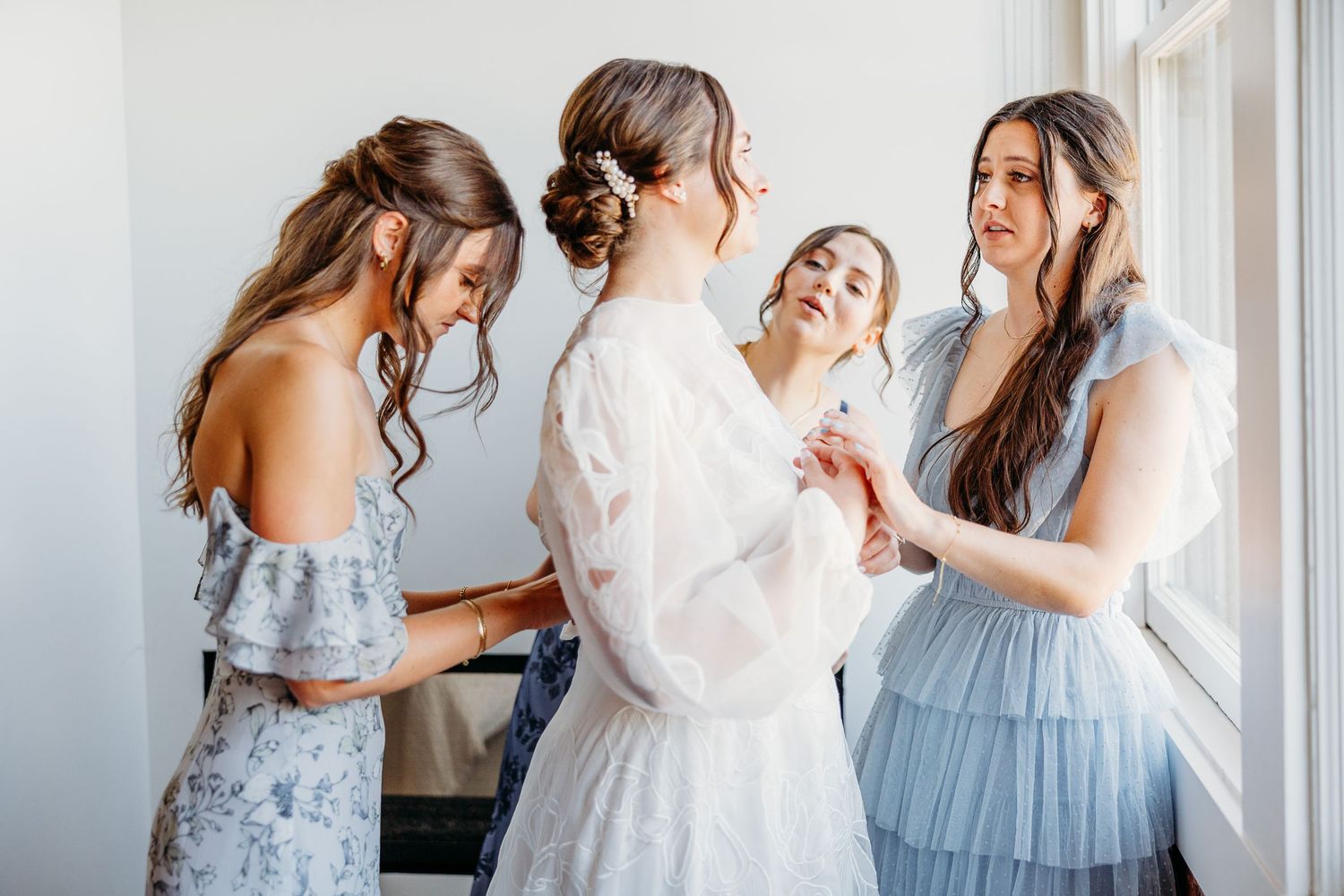 A group wearing soft blue bridesmaid dresses helps another in a white robe get ready by a bright window in a suite at Hotel Cerro SLO.