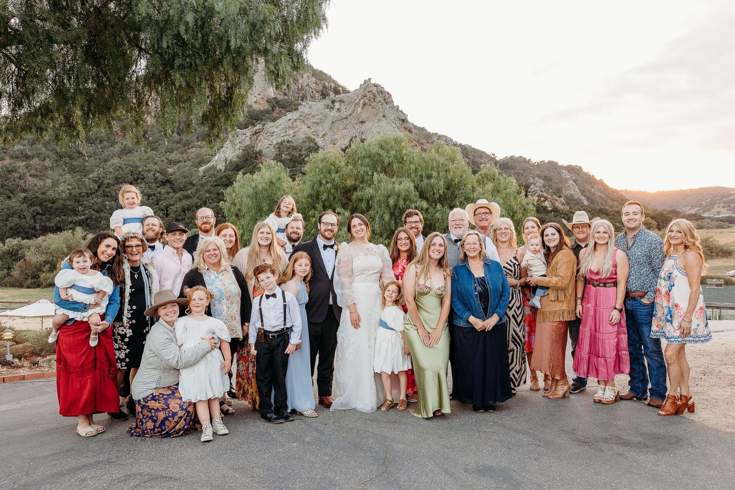Wedding party gathered outdoors for a group photo against a mountainous backdrop at sunset.