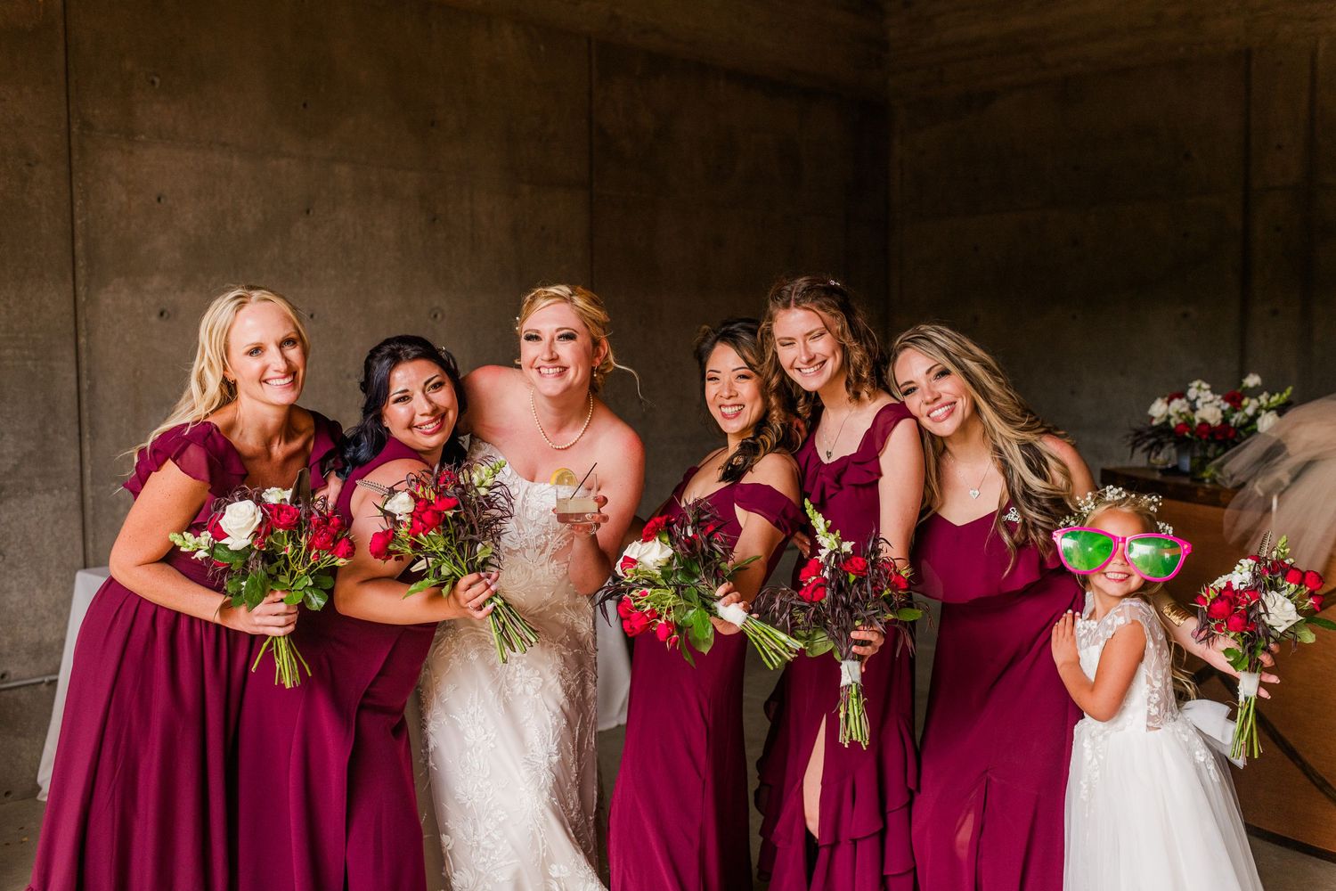 Bridesmaids in burgundy dresses pose with bride in lace gown holding red and white bouquets.