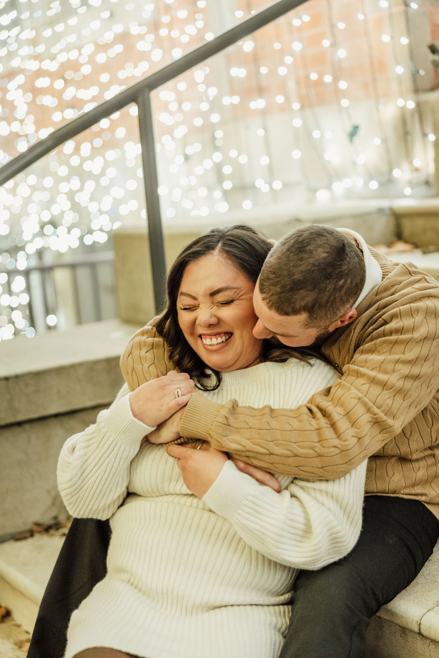 A couple in cozy sweaters shares intimate laughter while embracing on a bench with twinkling lights in the background.