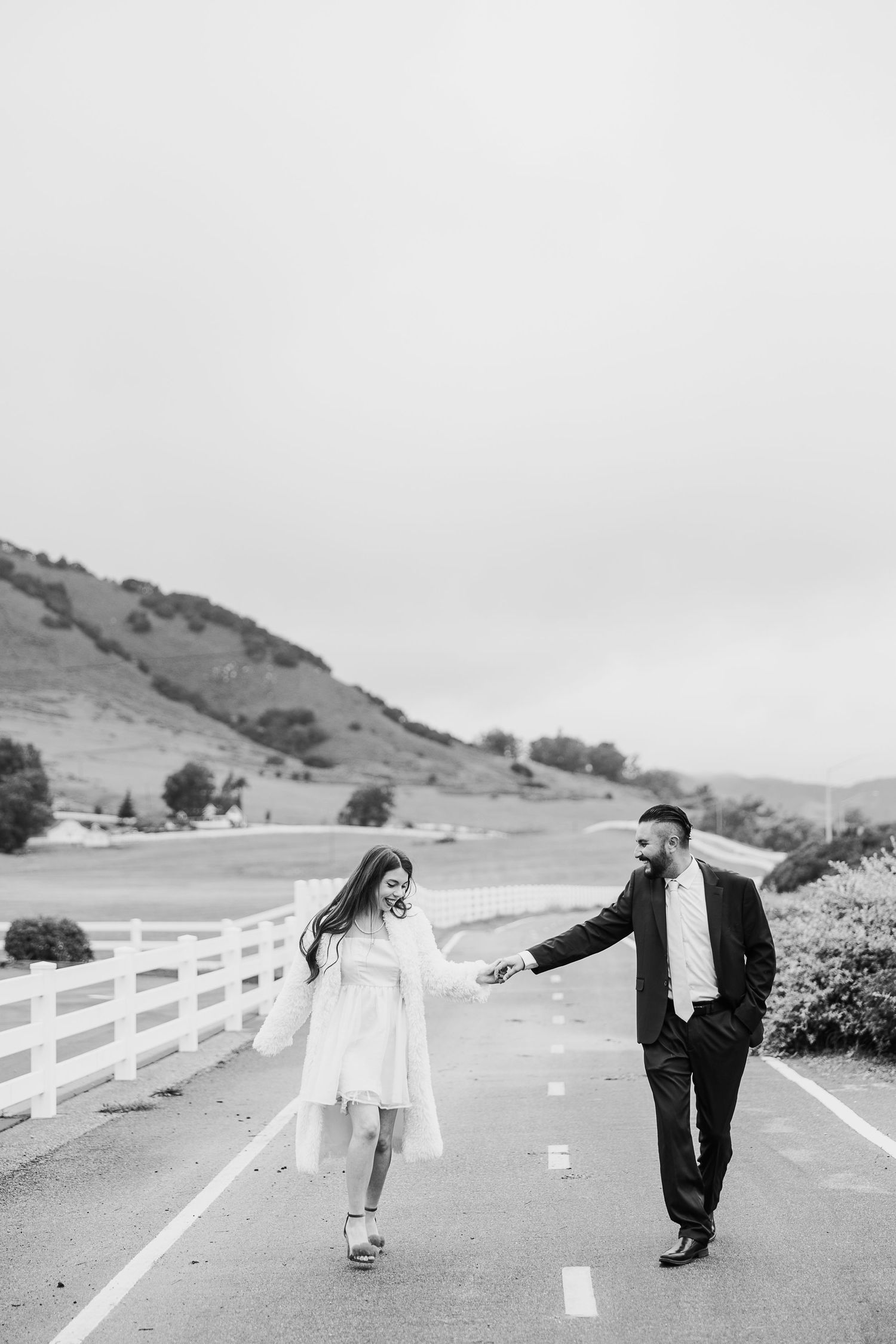 Two people walk together on a paved path alongside a white fence with rolling hills in the background.