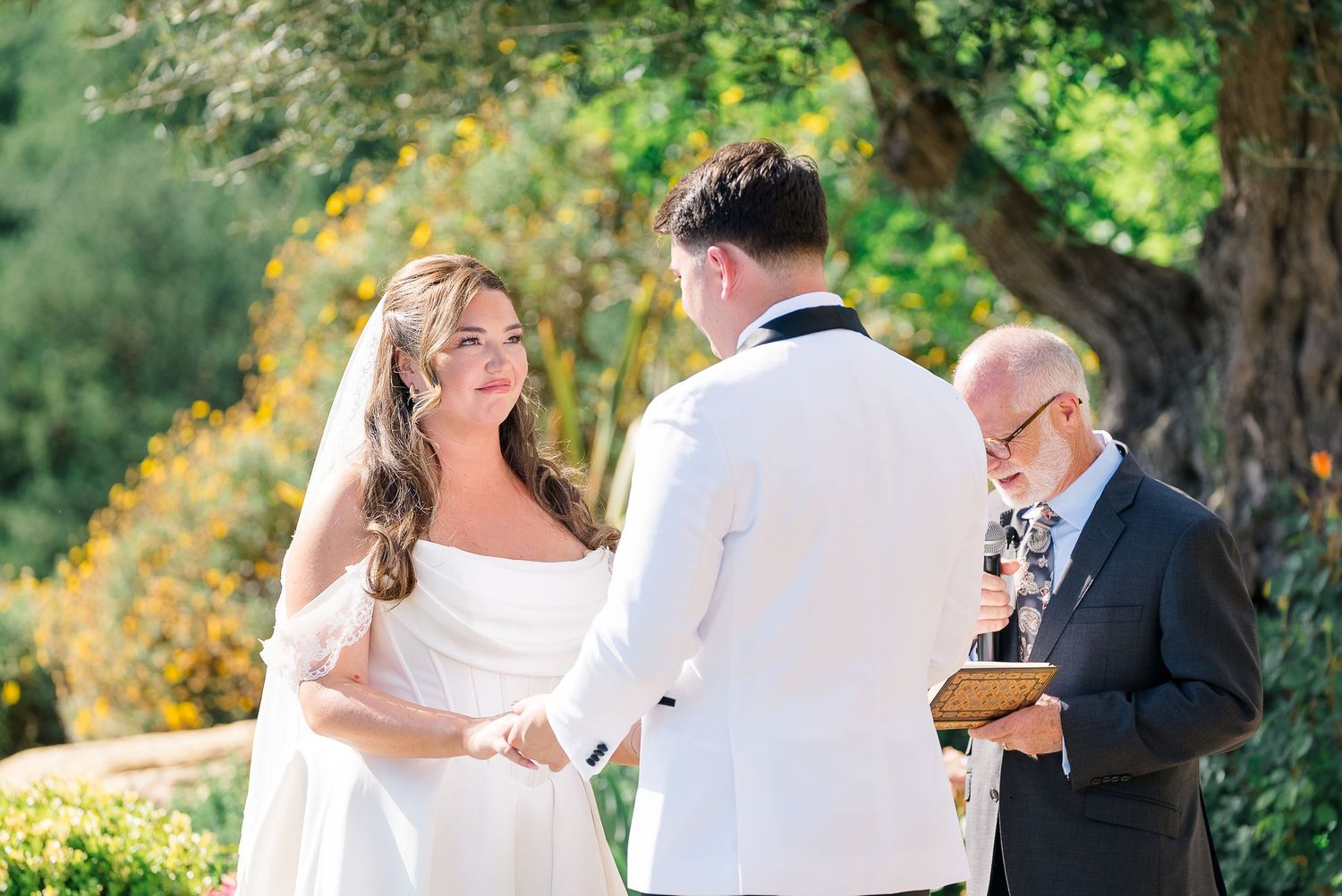 Bride and groom exchange vows outdoors with officiant during sunny garden wedding ceremony.