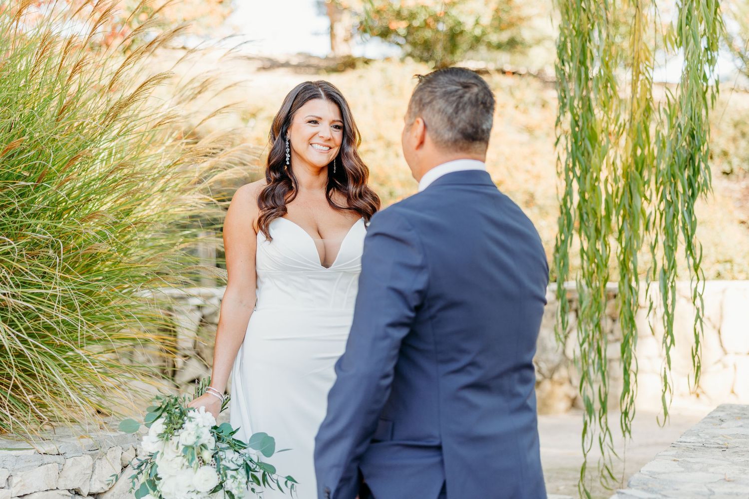 Bride in white gown smiles at groom in navy suit during outdoor first look ceremony with greenery backdrop.