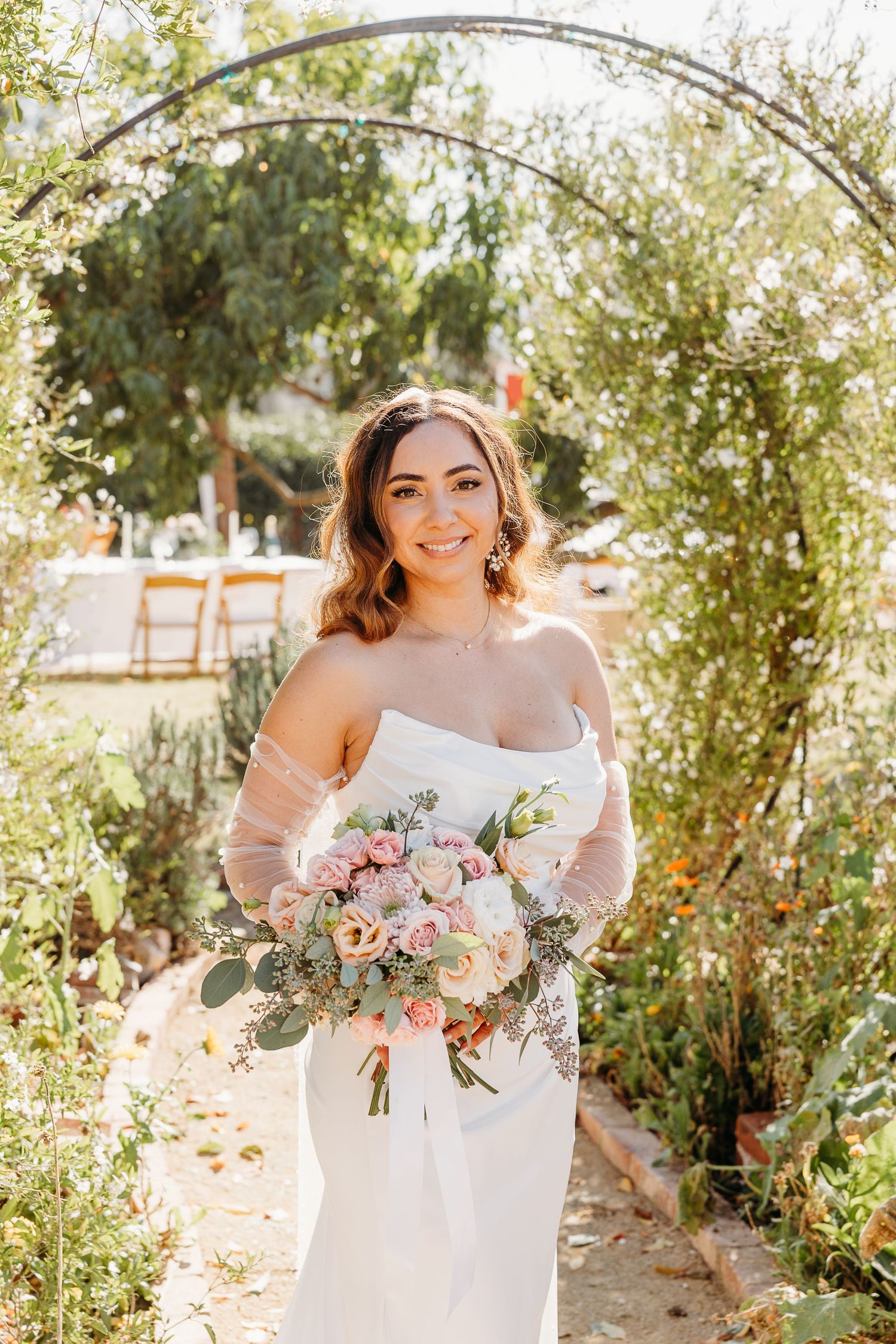 A bride in a strapless white wedding dress holds a pastel bouquet under a garden arch on a sunny day.