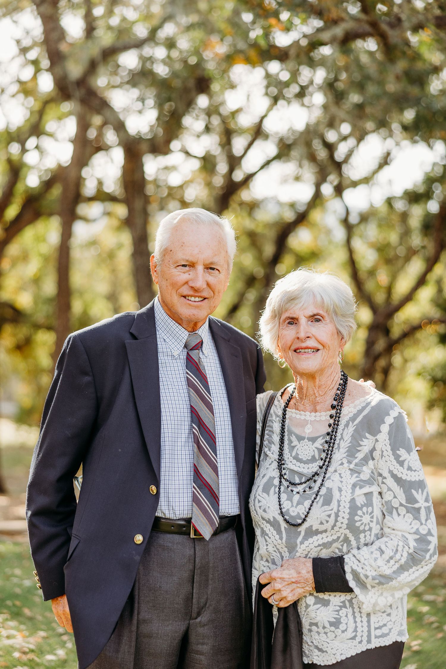Elderly couple poses together outdoors in formal attire with autumn trees in the background.