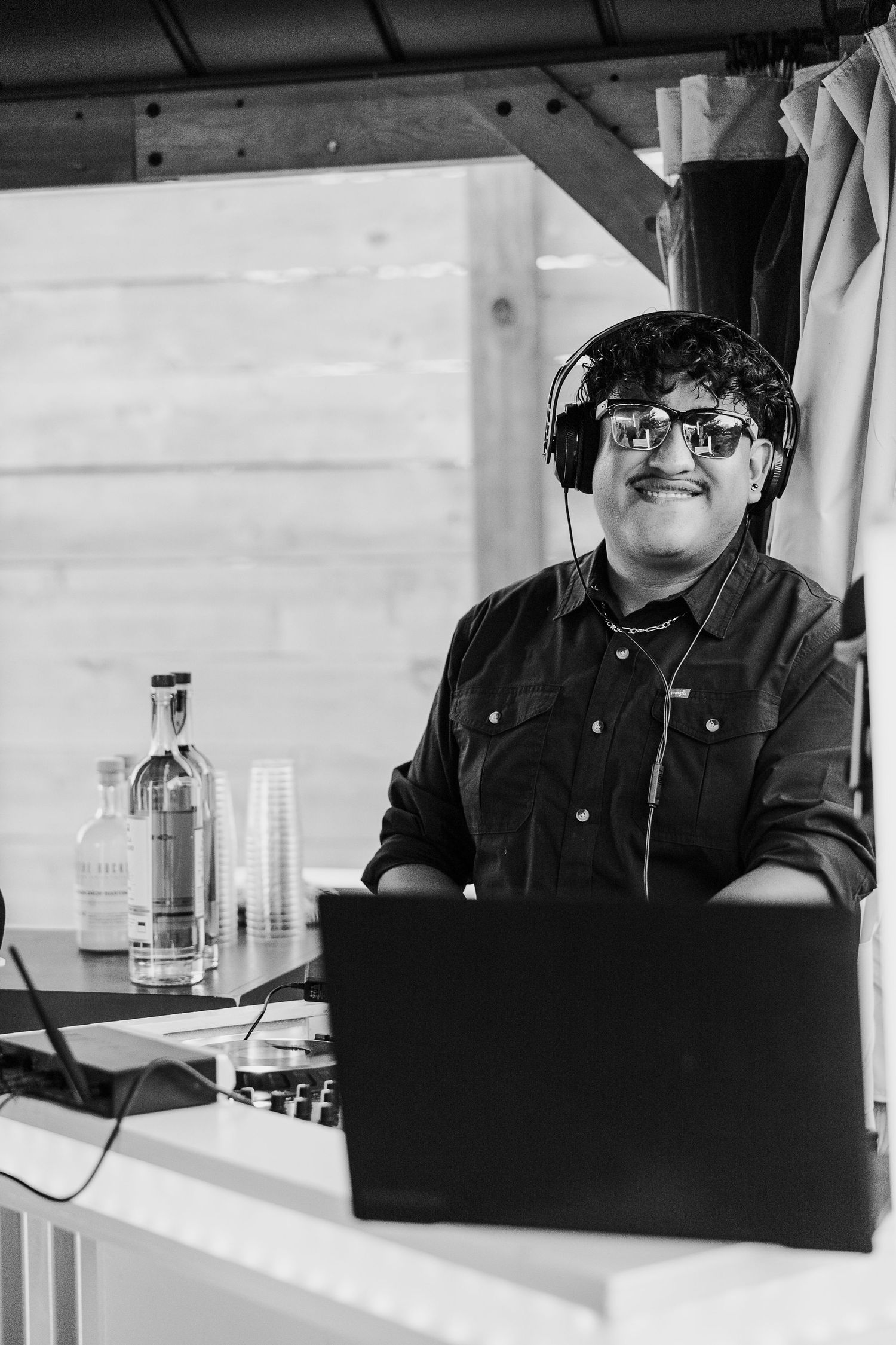 A DJ wearing headphones and glasses smiles while working at a setup with a laptop and liquor bottle in black and white.