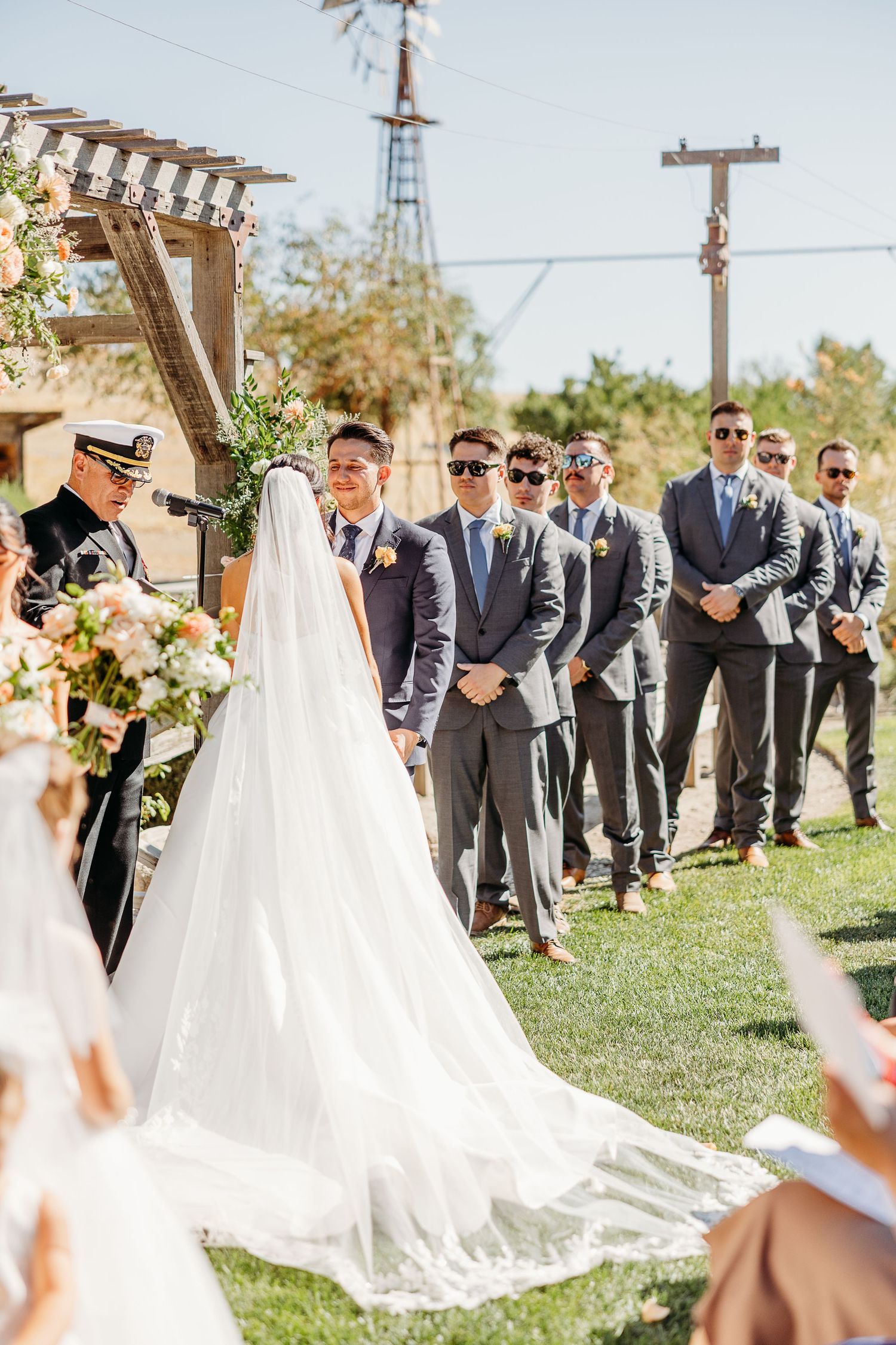 Wedding party in gray suits and white dresses gather outdoors under a pergola on a sunny day.