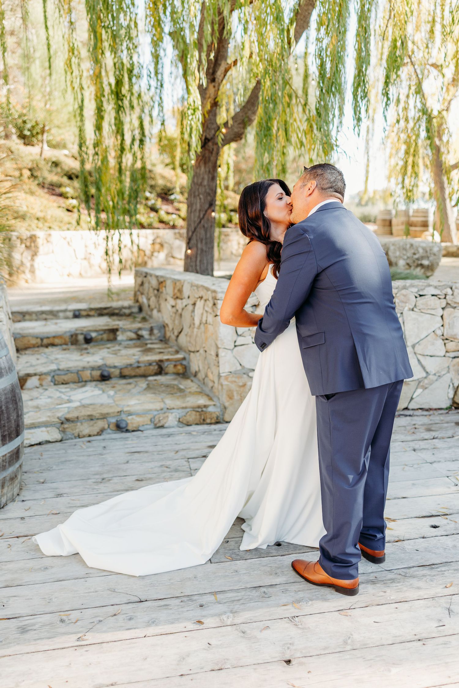 A romantic wedding moment captured under weeping willow trees on a stone patio.
