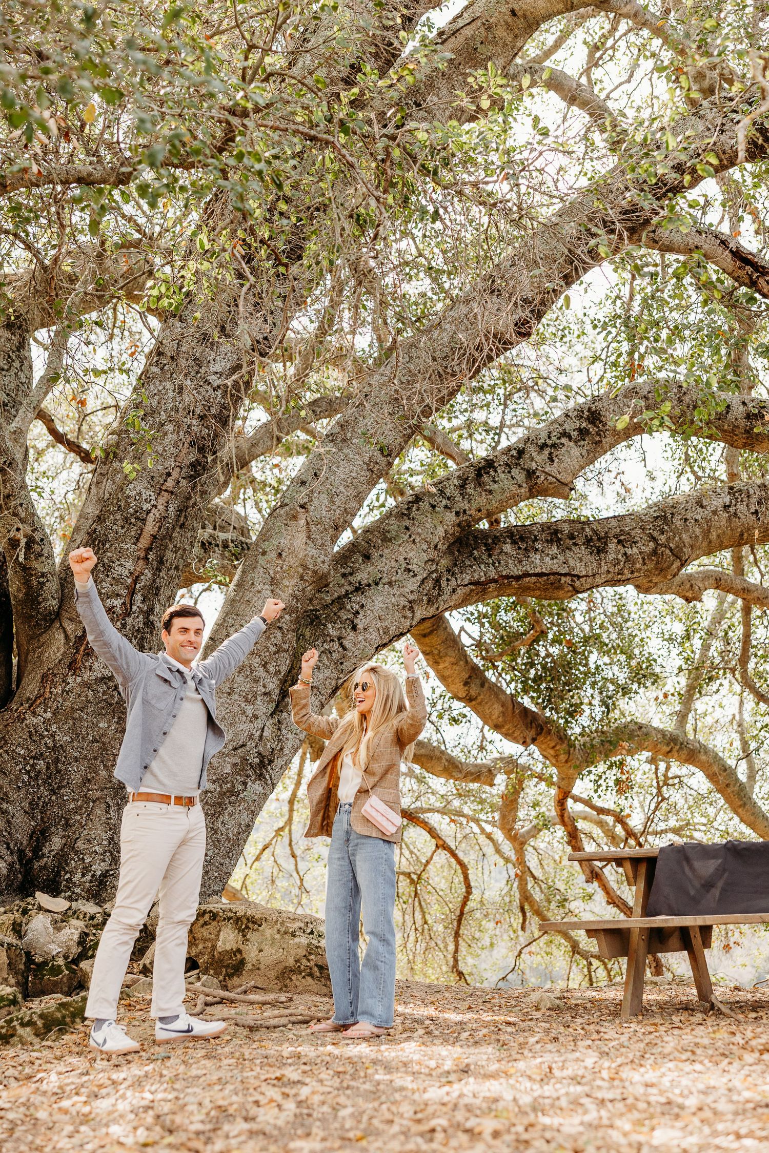 Playful couple dancing and twirling beneath sprawling oak tree branches in natural outdoor setting.