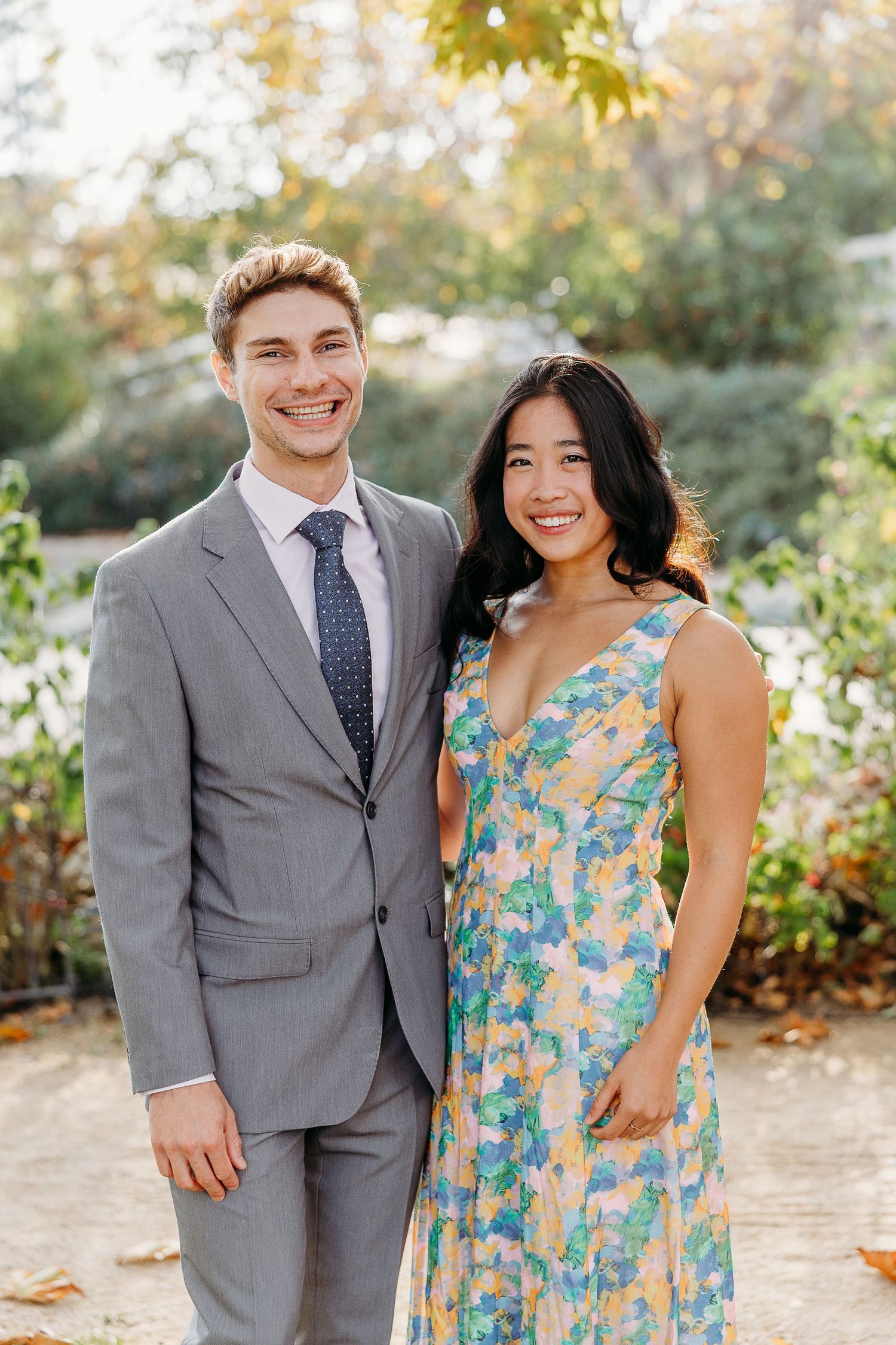 A couple poses outdoors in autumn, with the woman wearing a blue floral dress and the man in a grey suit.