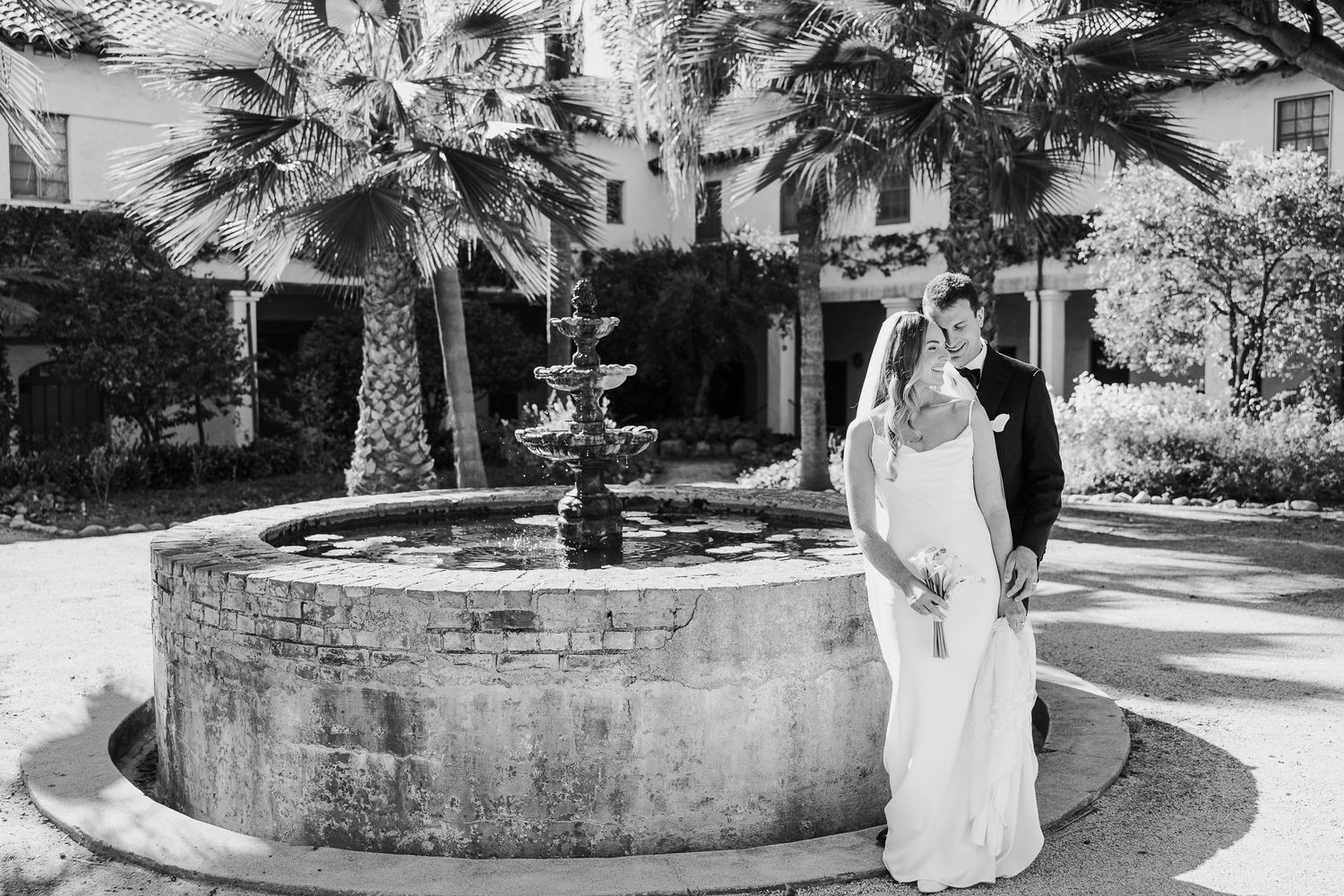 Elegant wedding photo session by a fountain surrounded by palm trees in a Mediterranean-style courtyard.