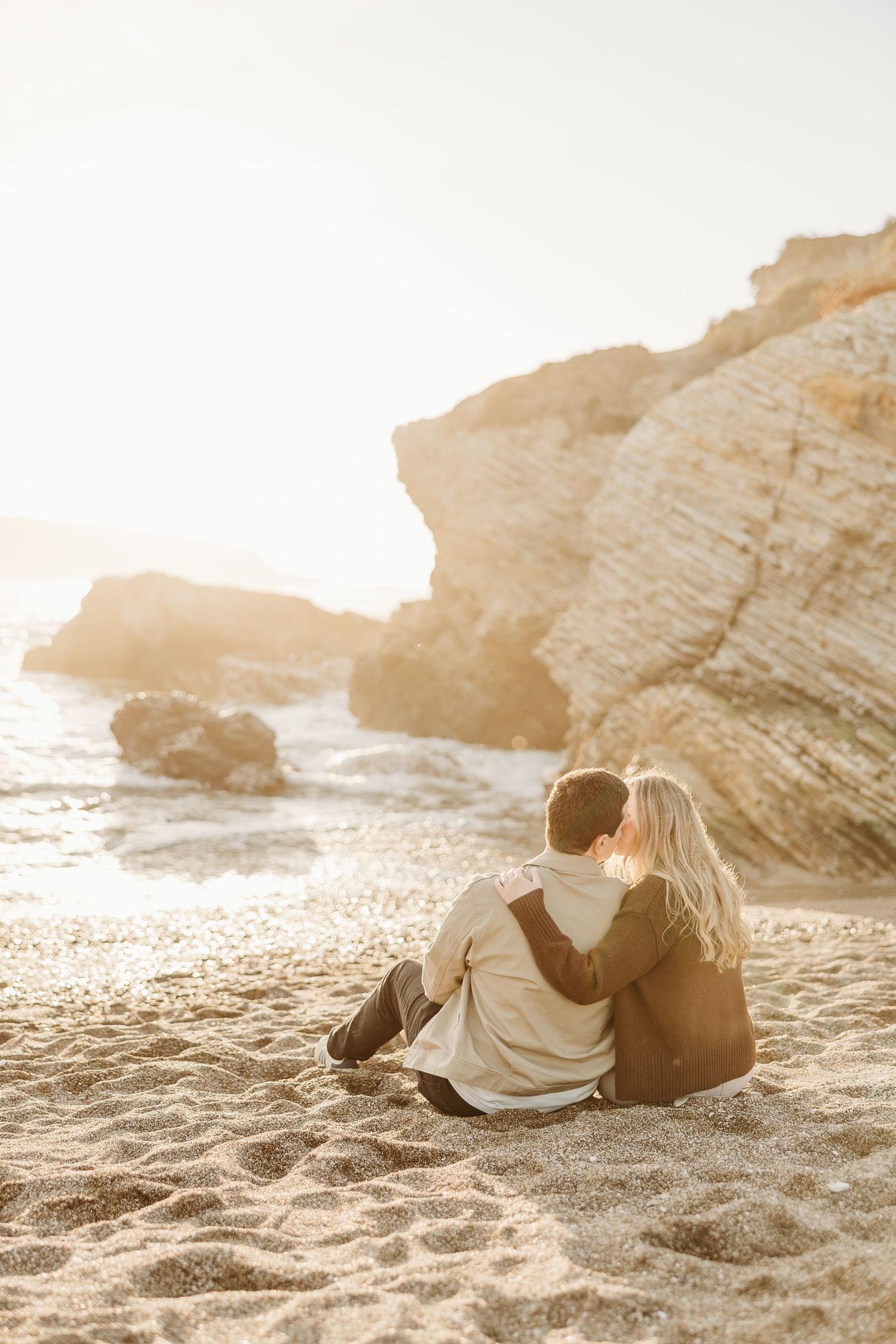 Couple lying on beach sand during golden hour with rocky cliffs in background.
