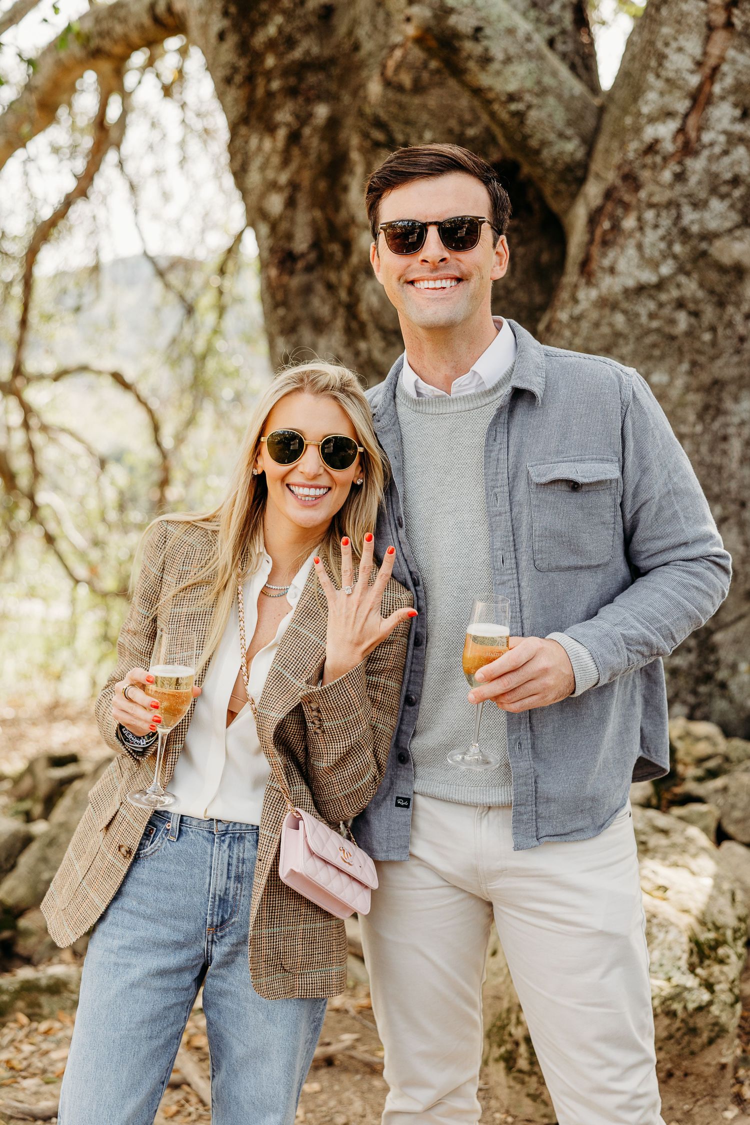 A couple wearing sunglasses and casual attire hold drinks outdoors while showing off an engagement ring.
