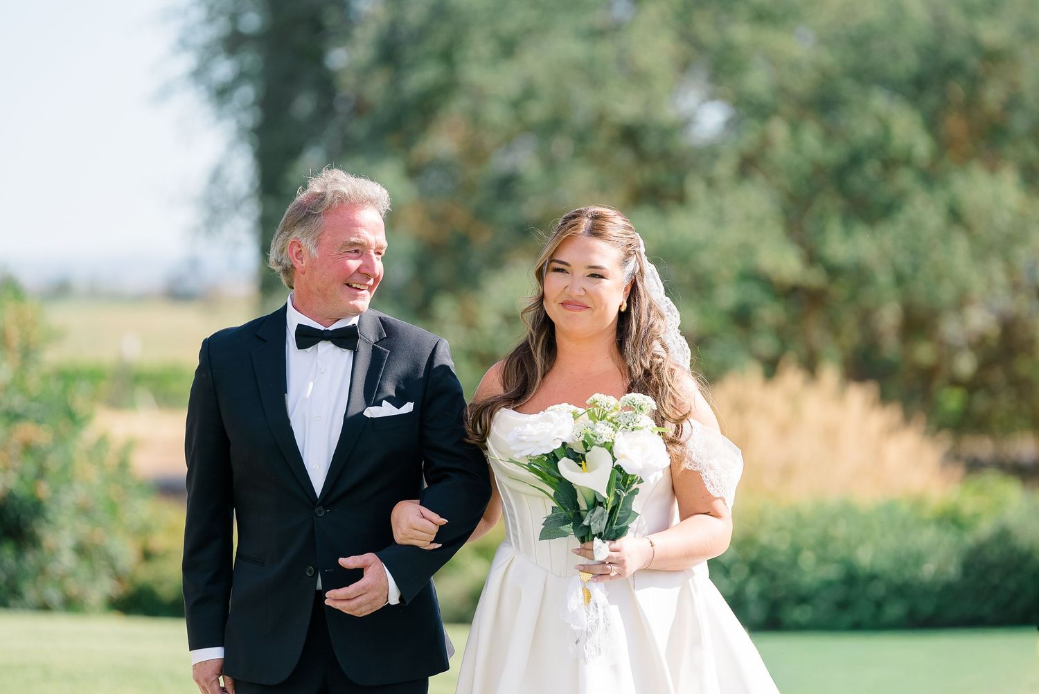 Bride in white gown holding bouquet walks with man in tuxedo outdoors at sunny wedding ceremony.