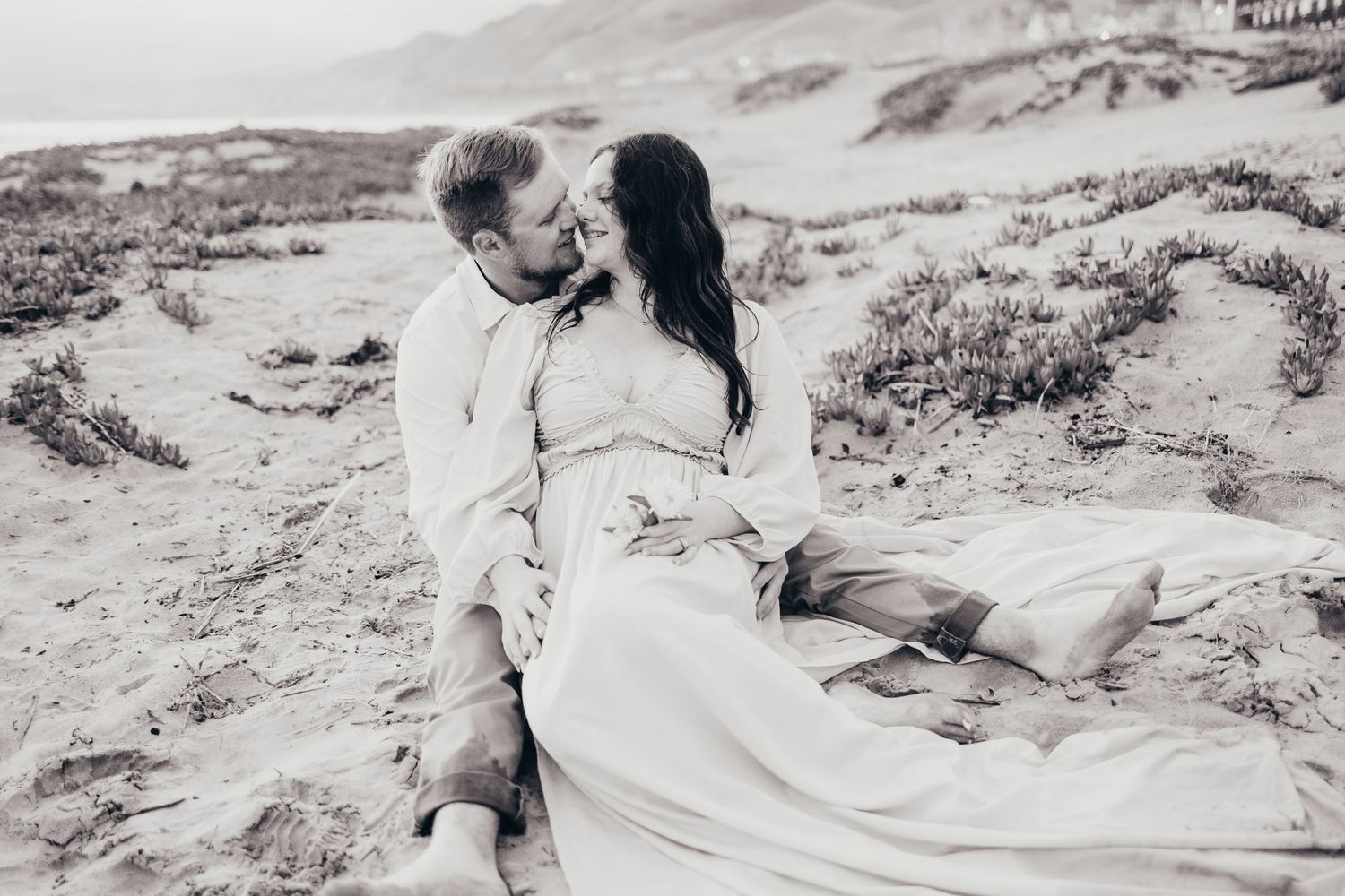 A black and white photo captures a tender embrace on a coastal desert landscape.