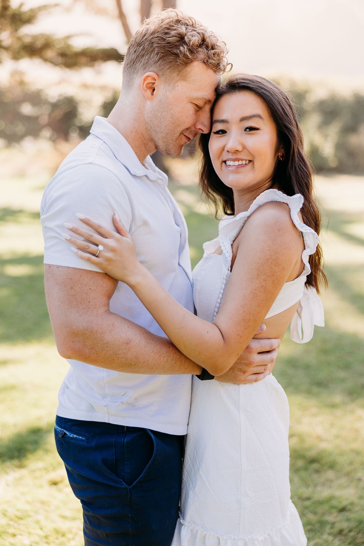 Couple in coordinated white outfits embracing in a sun-drenched park setting.