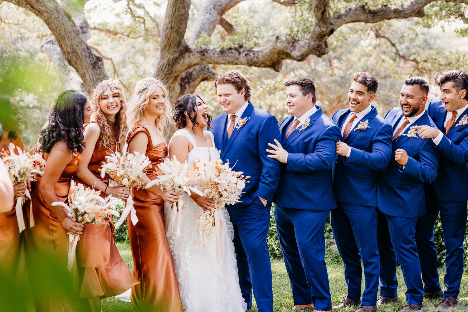 Wedding party in blue suits and copper dresses posing together under trees.