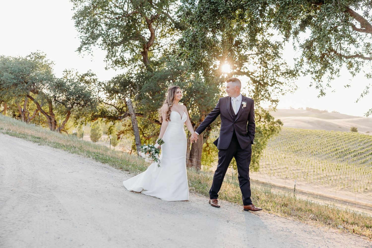Wedding couple walks hand in hand through scenic countryside setting at sunset.