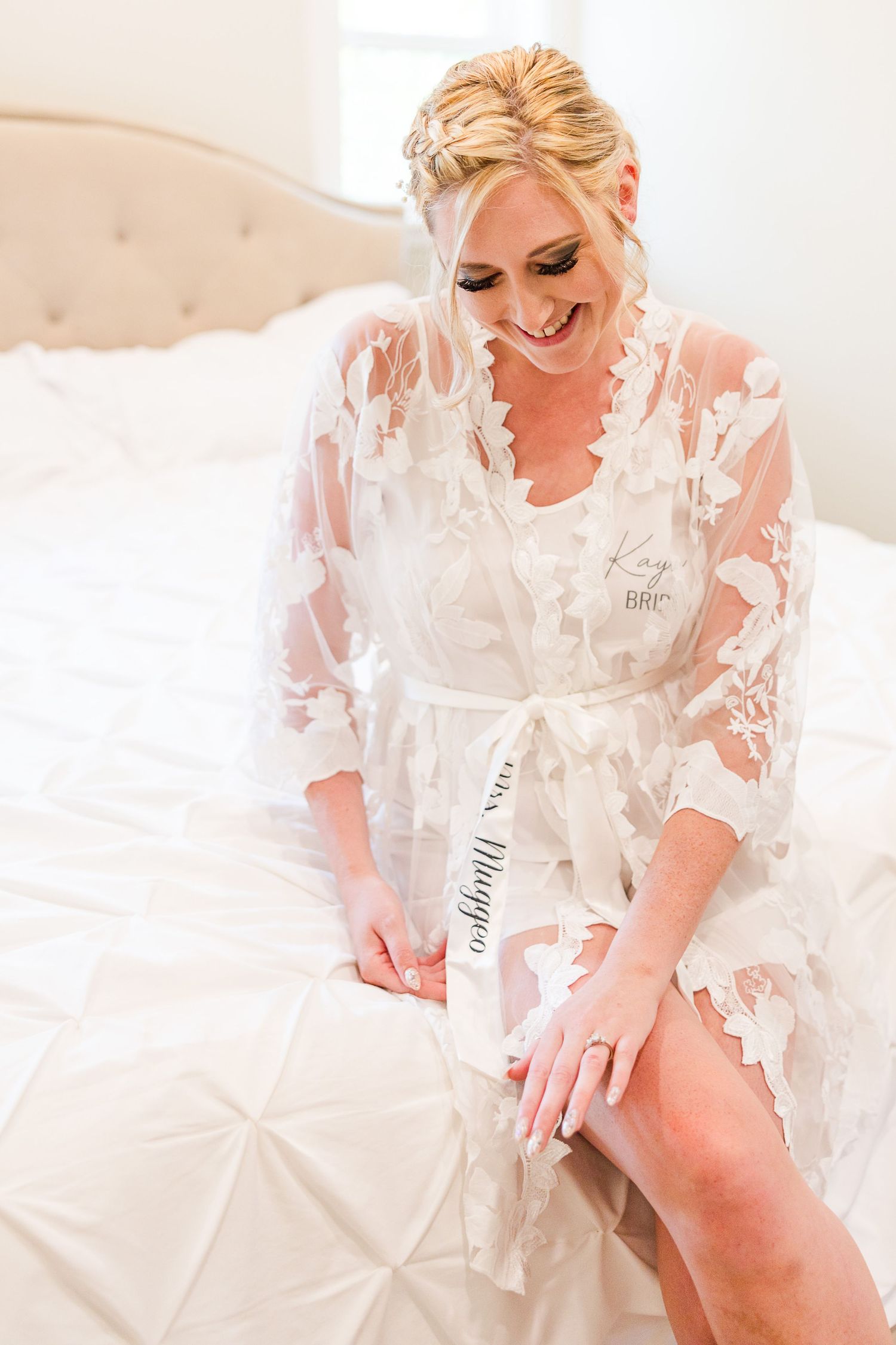 Person in white lace robe sits on plush bed during wedding morning preparations.