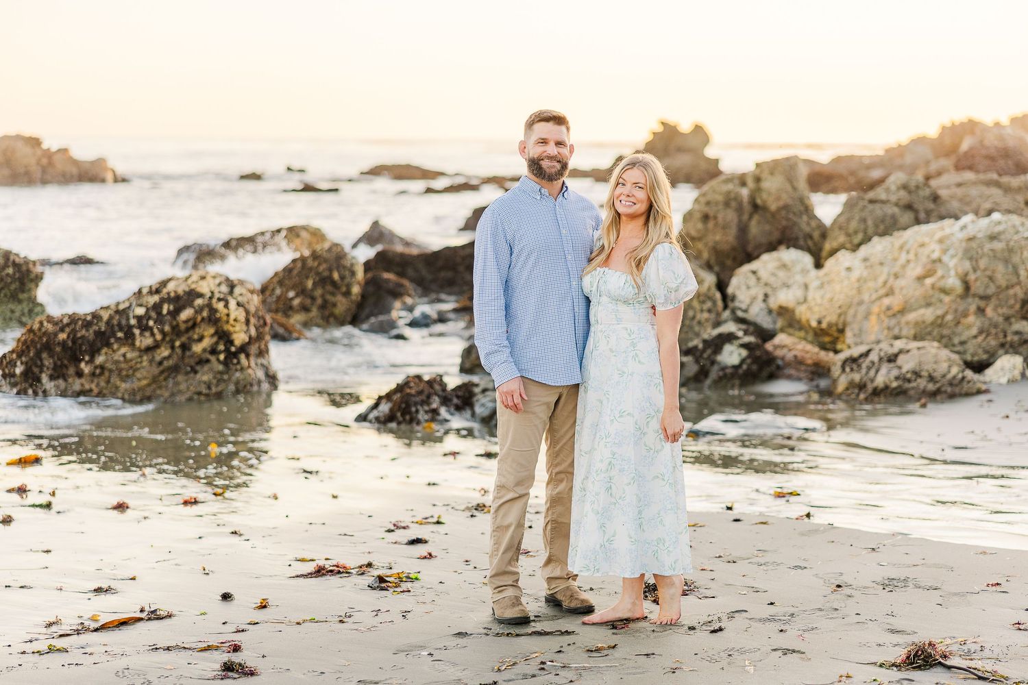 Romantic couple strolls along rocky beach coastline at sunset creating silhouettes against golden sky.