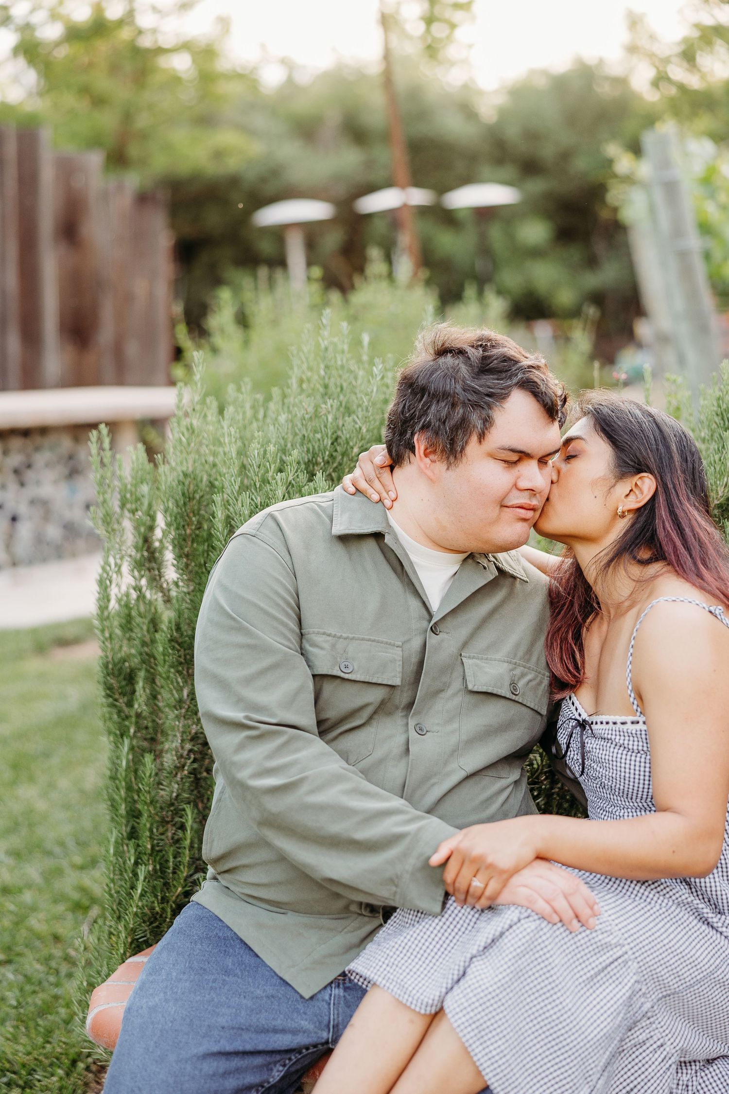 A romantic outdoor portrait in summer featuring a couple sharing an intimate moment on a bench by rosemary plants.