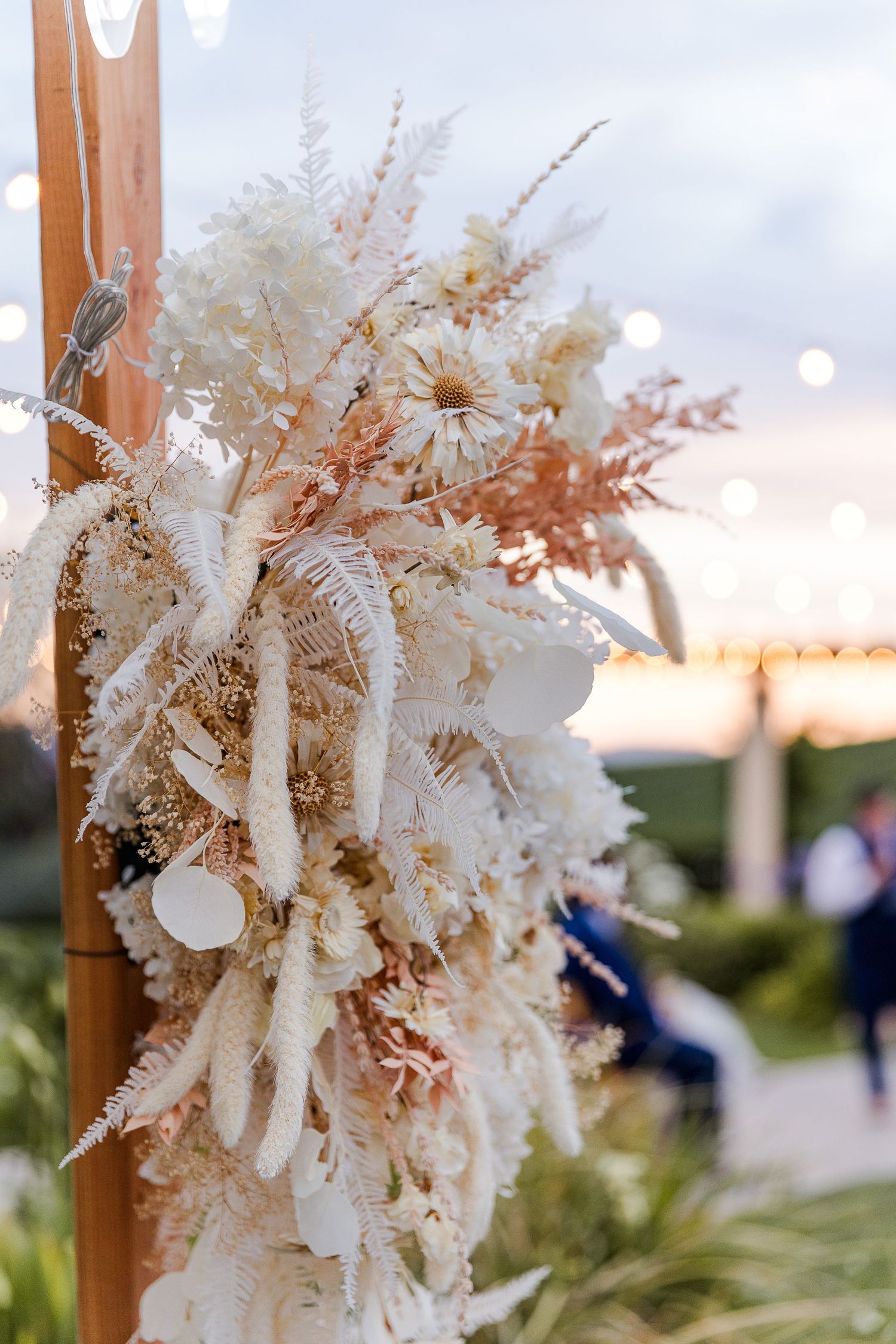 Dried pampas grass and floral arrangement adorns wooden wedding arch structure.