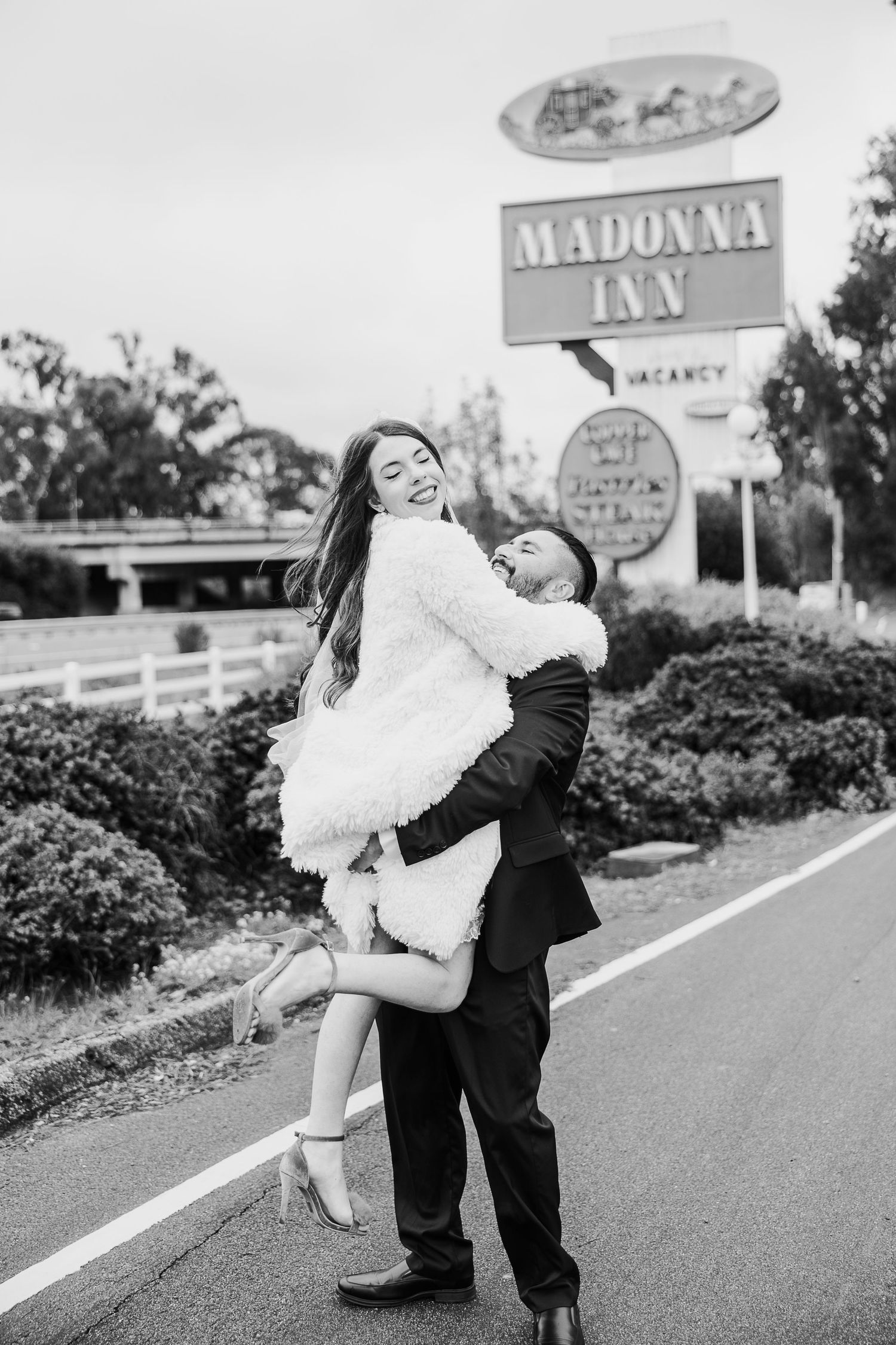 A couple in formal wedding attire embrace playfully on a roadway with vintage motel signage visible in the background.