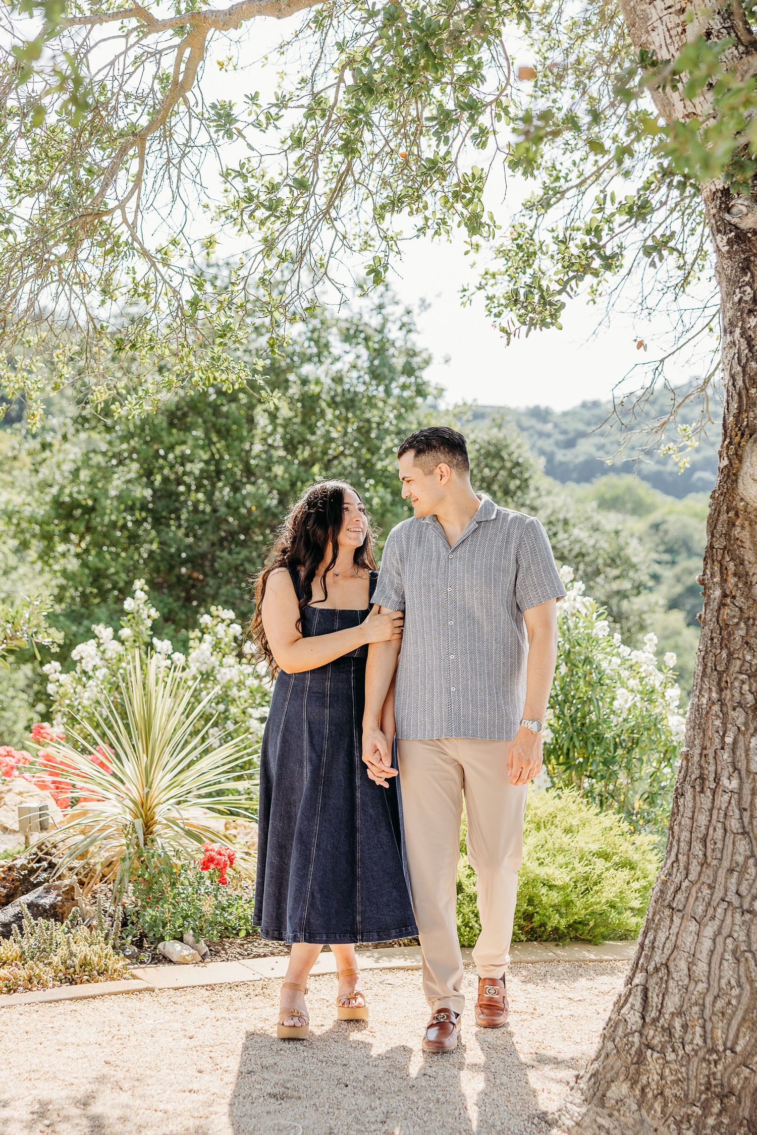 Two people walking together on a scenic path surrounded by trees and desert plants in a natural outdoor setting.