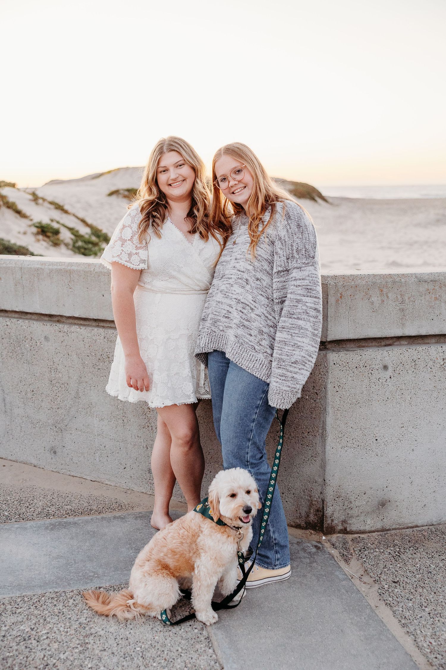 Two people in casual attire pose with a small white dog on a beach walkway at sunset.