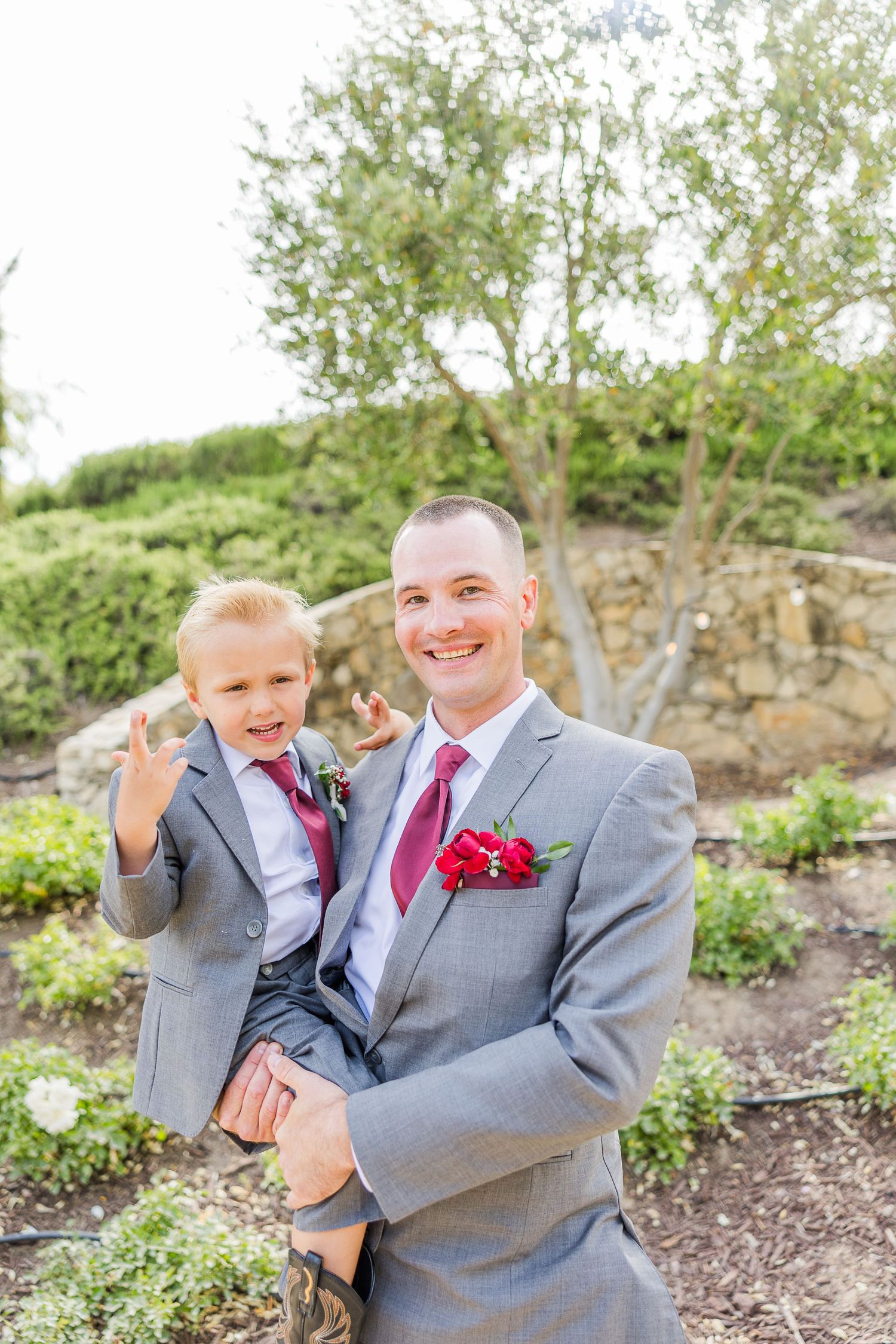 Wedding day portrait in grey suit with red boutonniere against garden hillside.