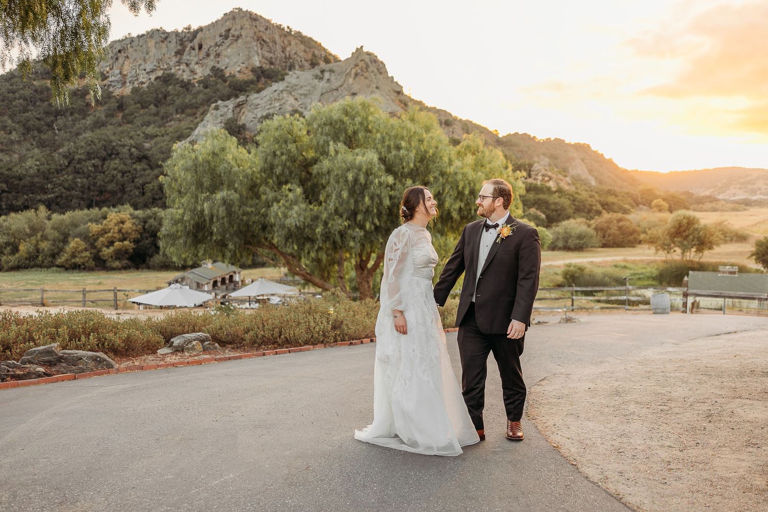 Newlyweds stand together on a path with scenic mountain backdrop at golden hour.