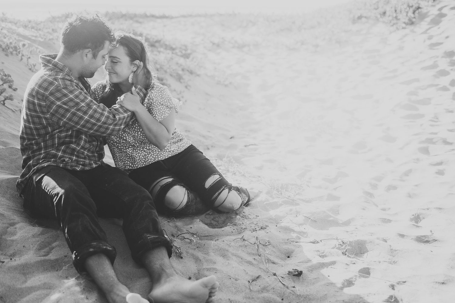 Couple sitting close together on sandy beach in black and white photo.