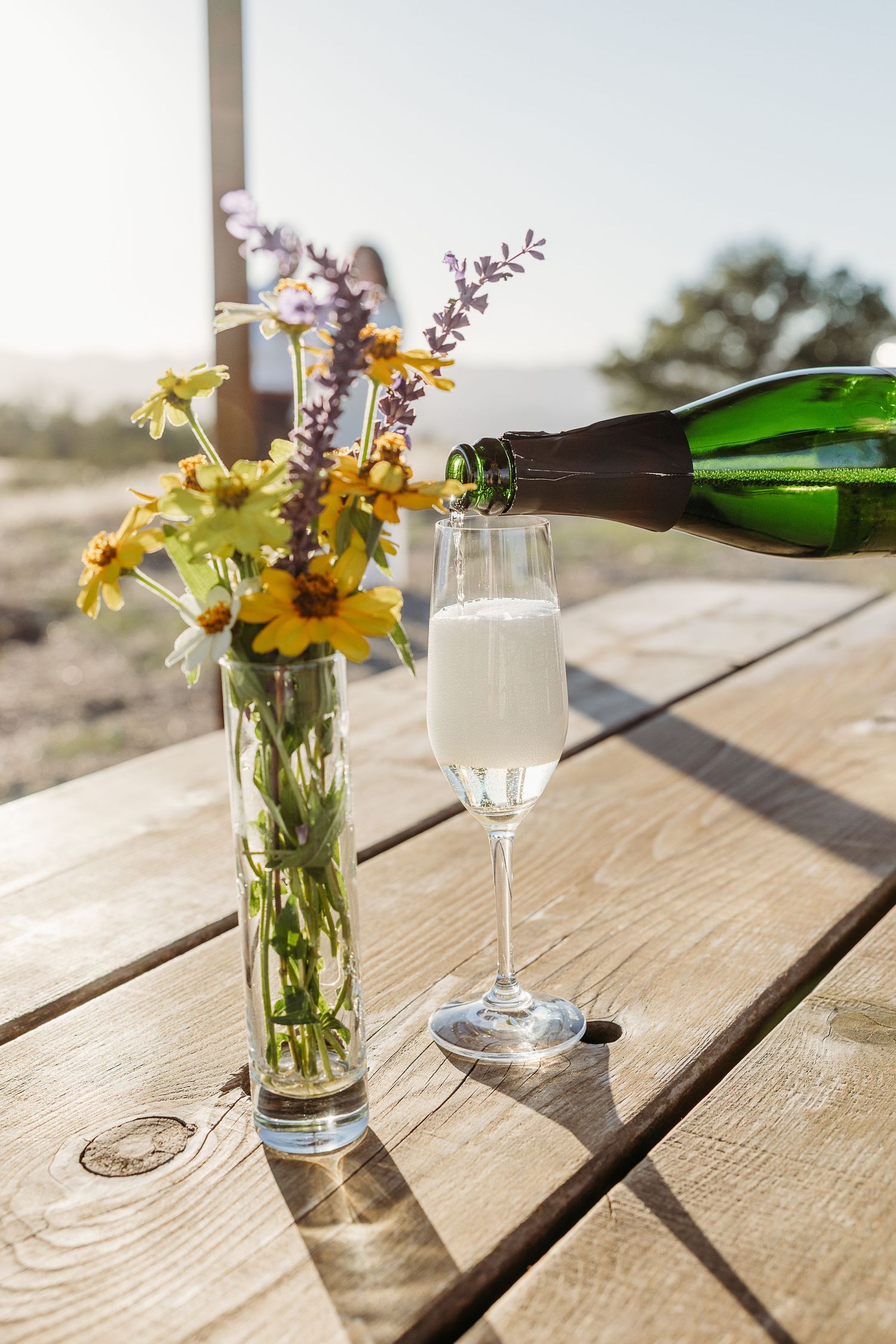 A series of photos showing champagne being poured into a glass next to yellow wildflowers in a vase on a wooden table.