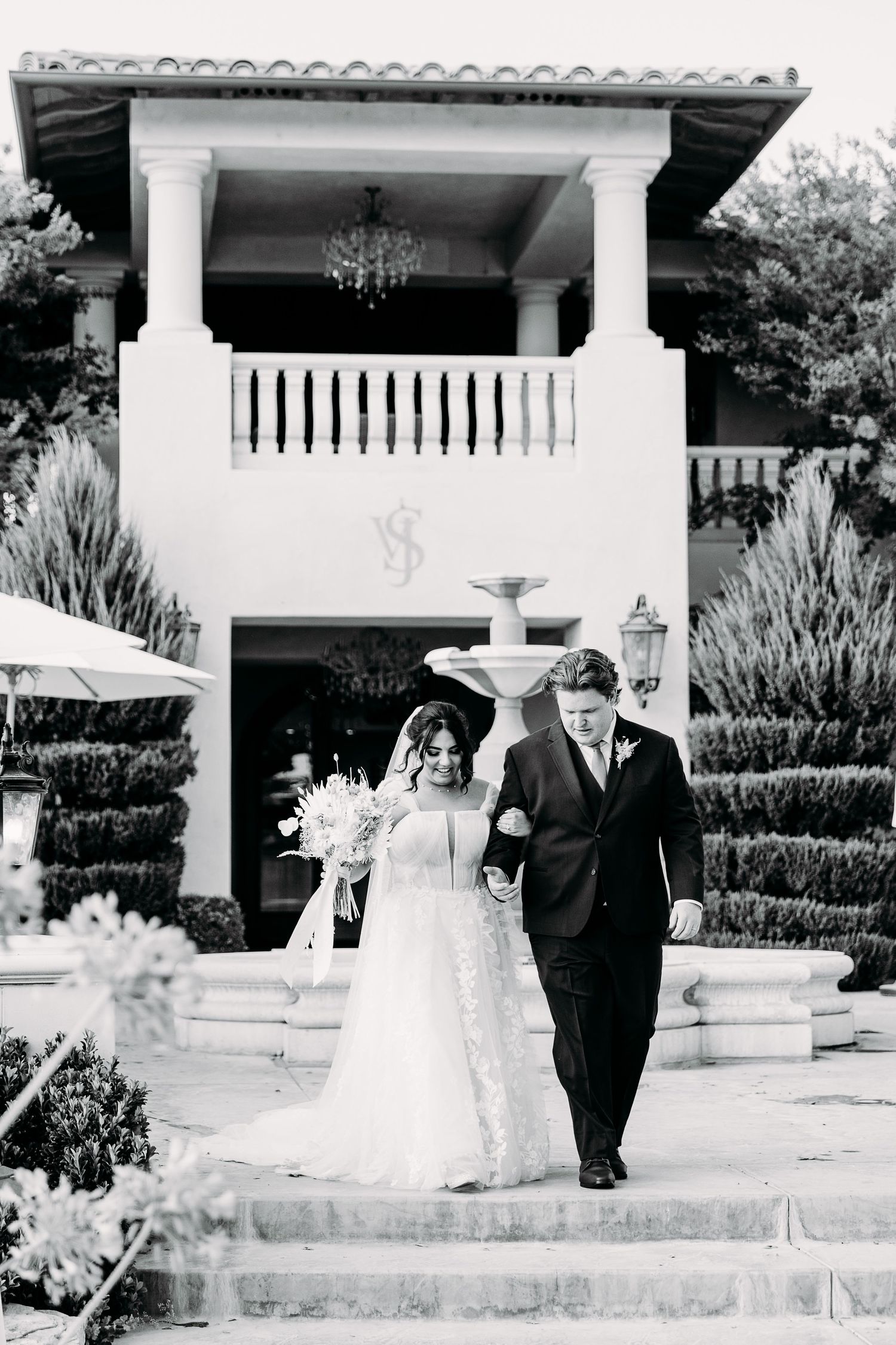 Black and white photo of couple walking down steps at Mediterranean style venue with fountain.