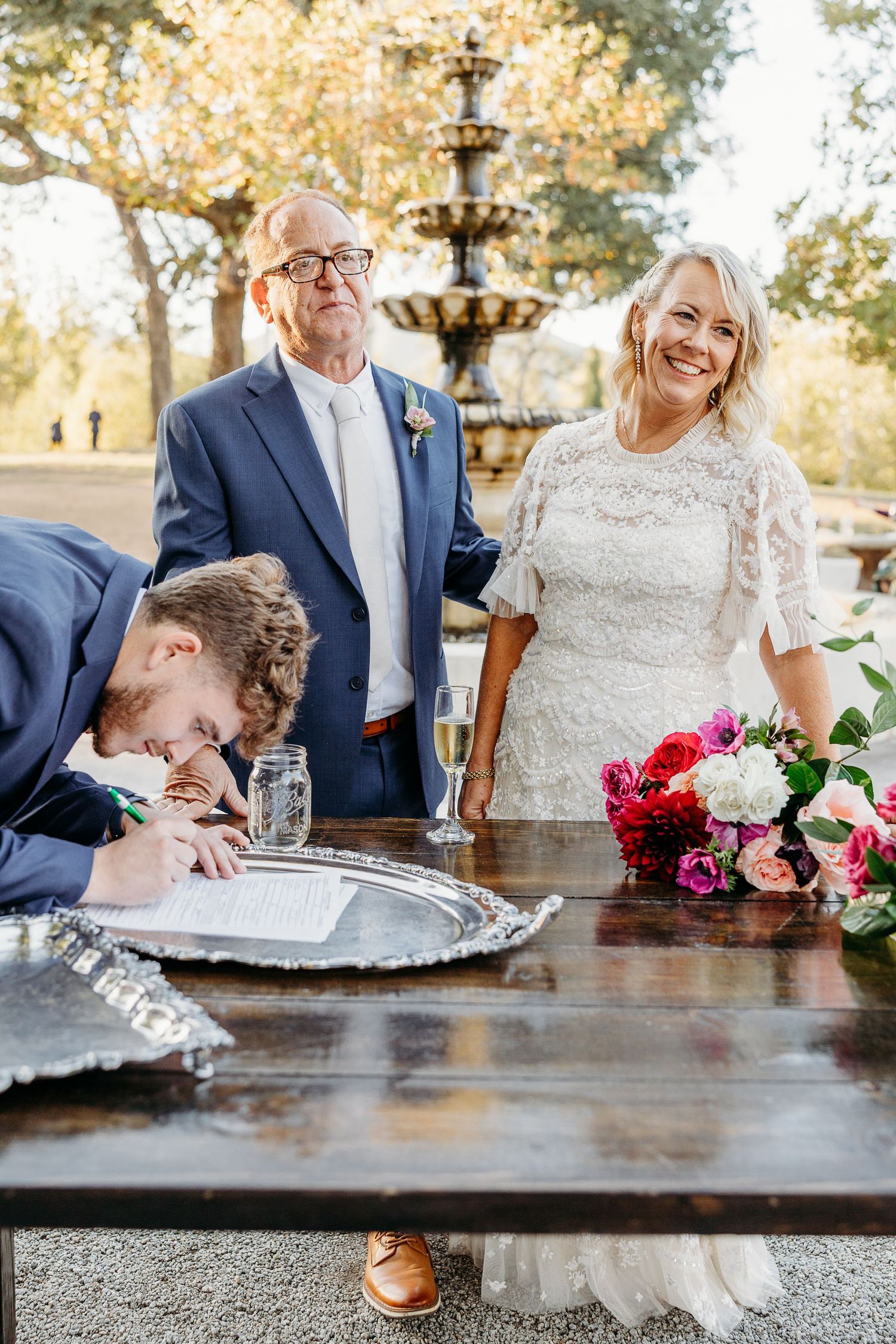 Wedding party signing documents at a dark wooden table with floral decorations.