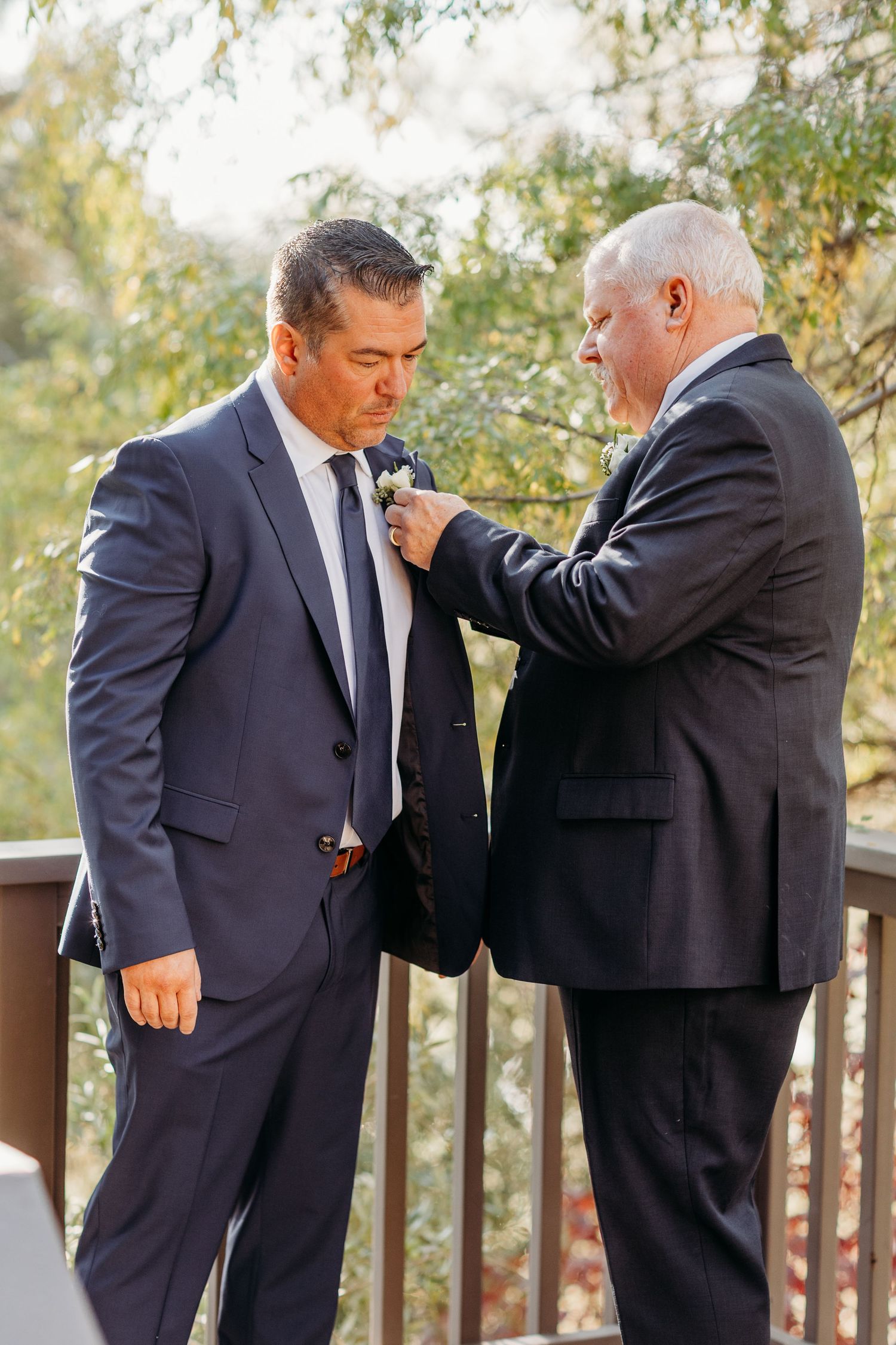 Two men in suits adjusting formal attire outdoors with trees in the background.