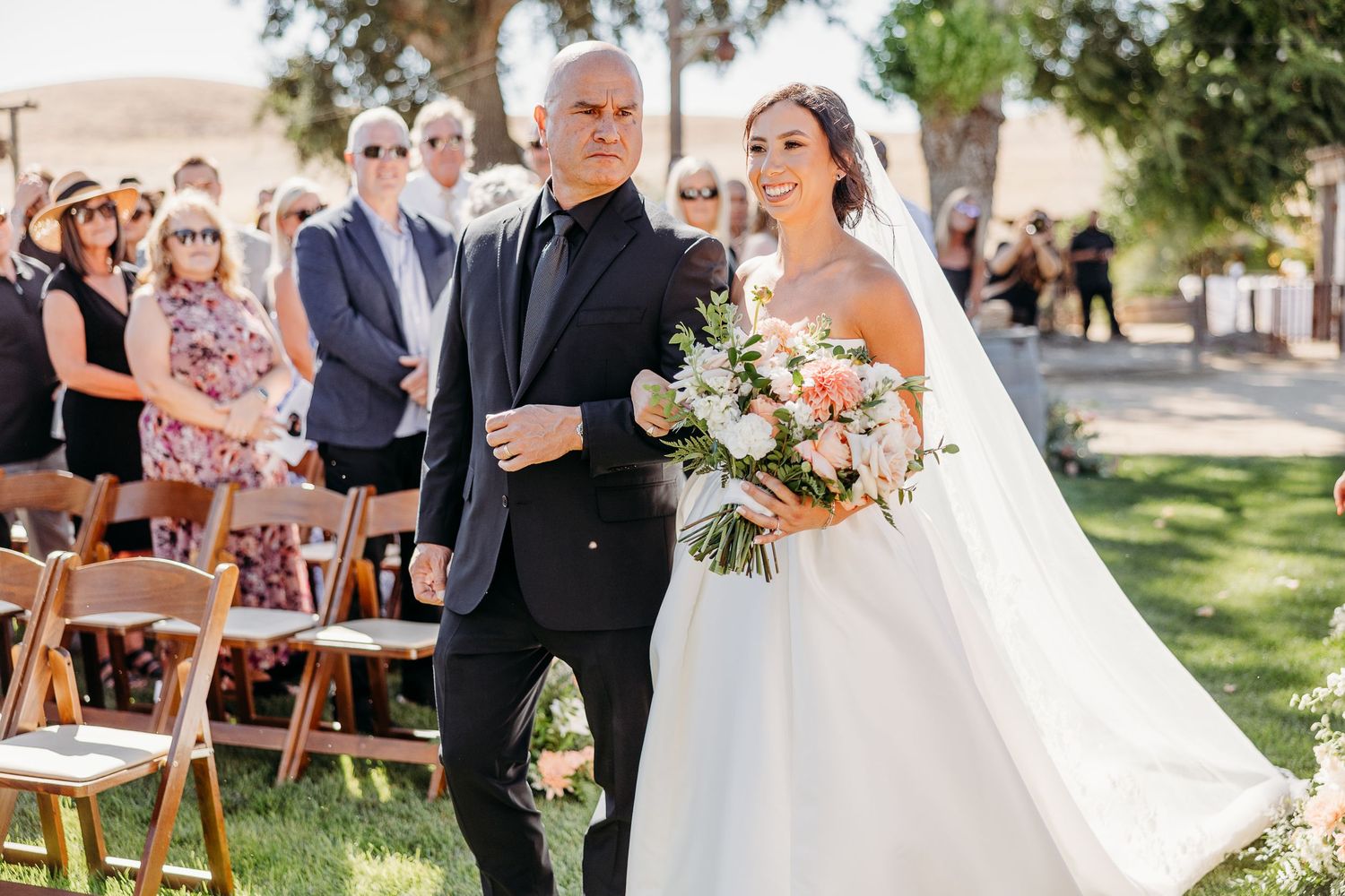 A joyful outdoor wedding ceremony with guests seated on wooden chairs under sunlit trees.