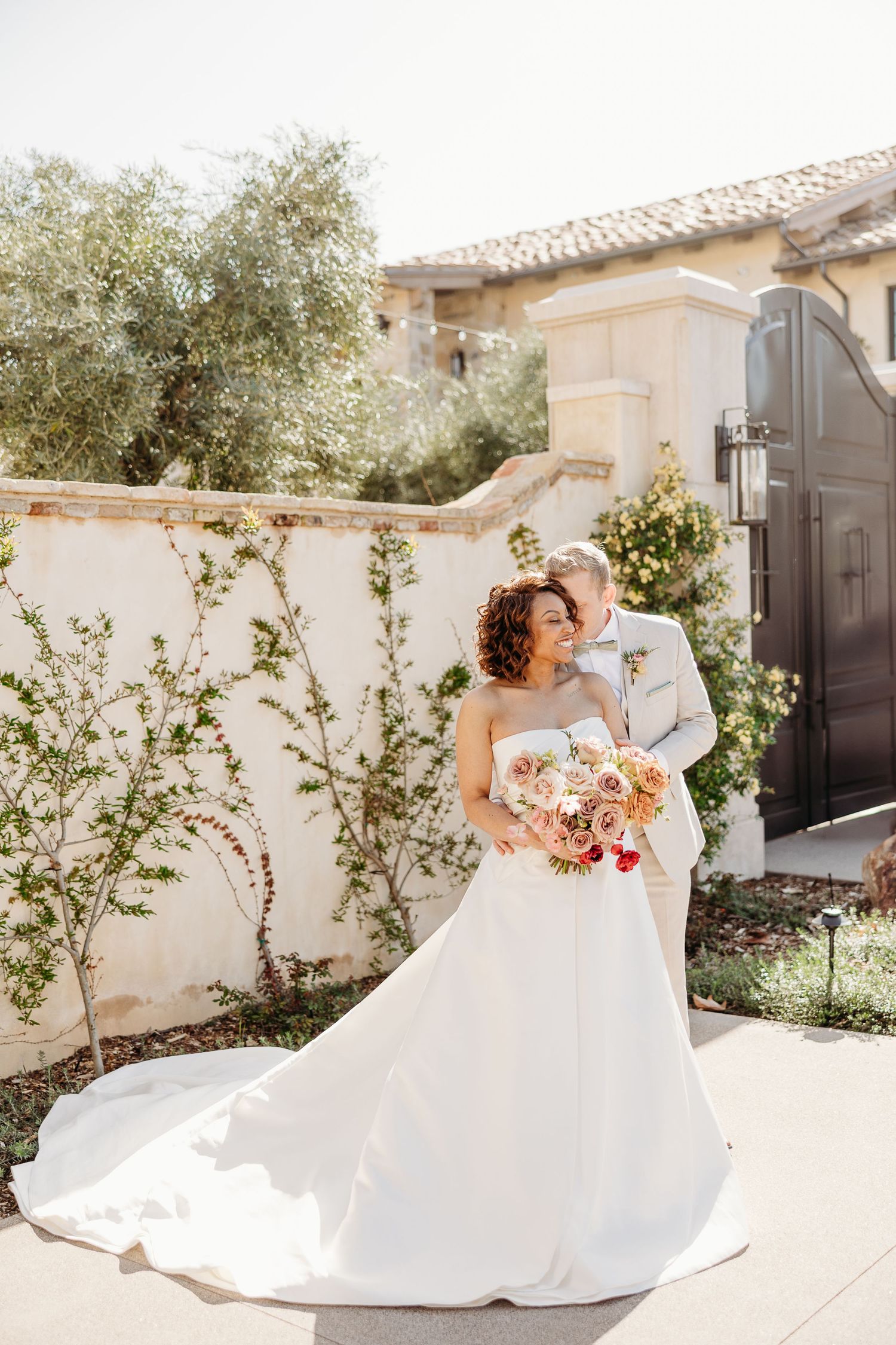 A romantic wedding couple embraces in a sun-drenched courtyard with Mediterranean architecture and lush greenery.